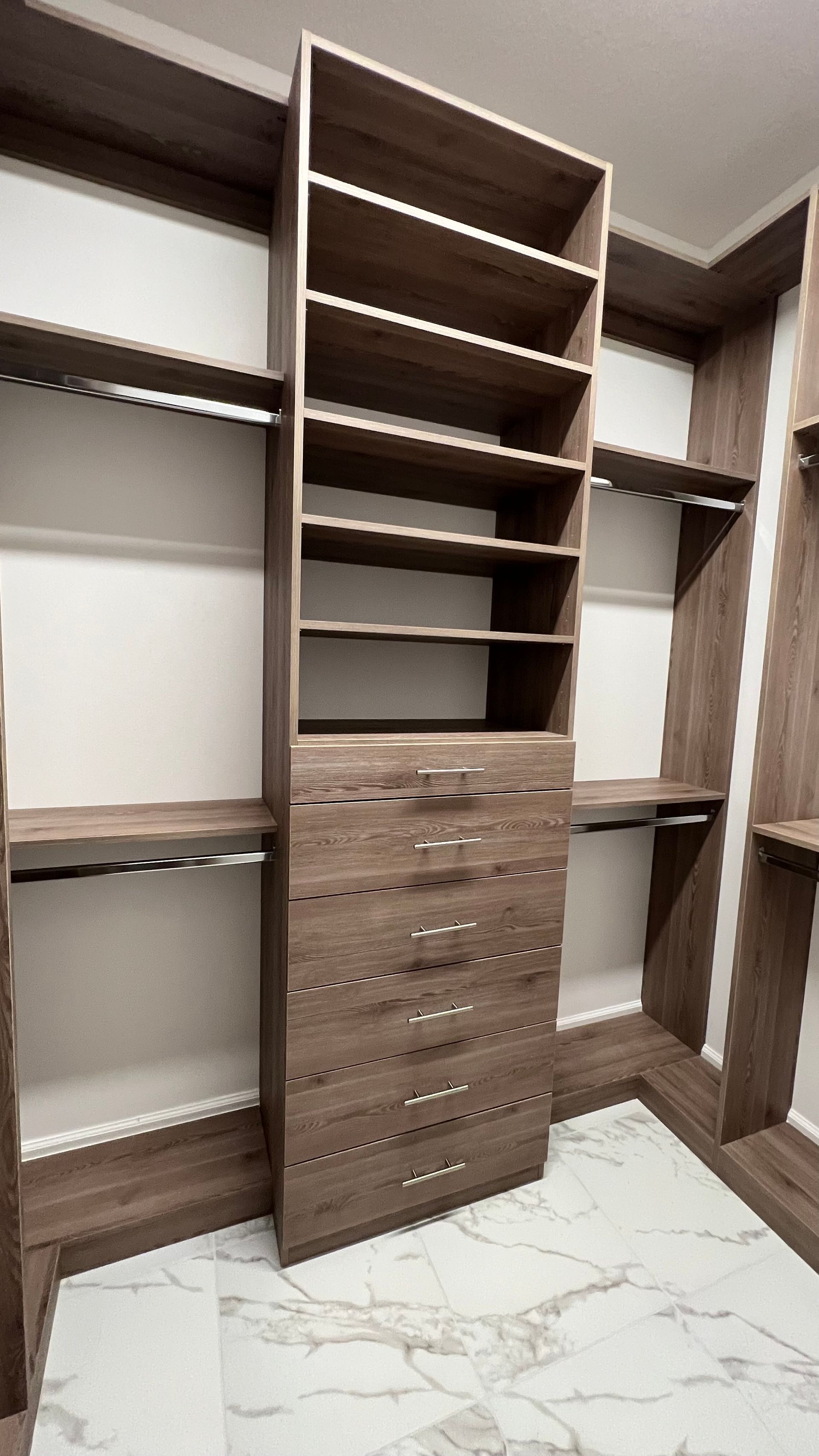 A walk-in closet featuring wood-tone shelves and a central drawer stack, set against white walls and a marbled floor.