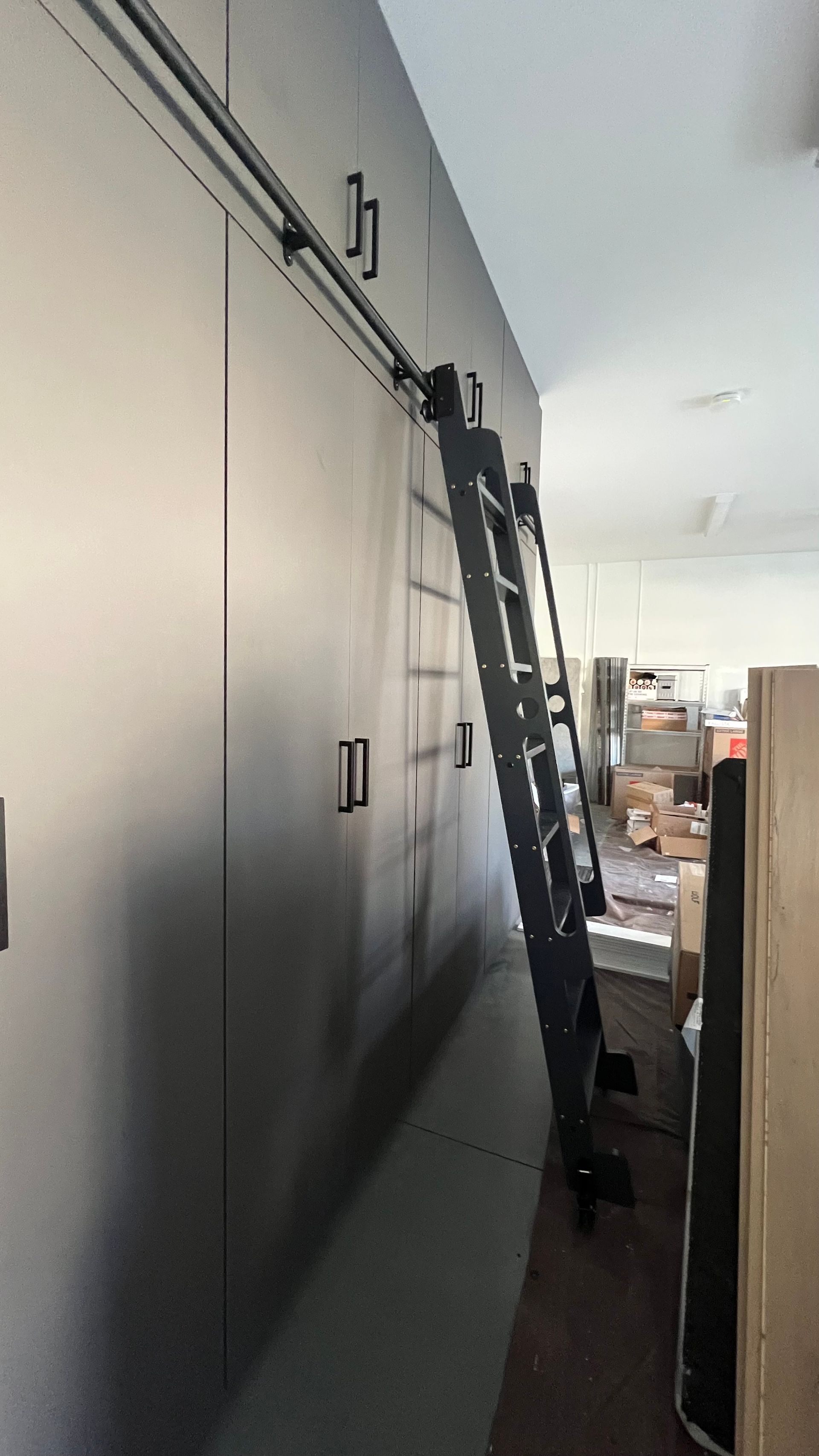 A tall, black rolling library ladder attached to a ceiling-mounted track against a wall of matte-finish cabinetry.