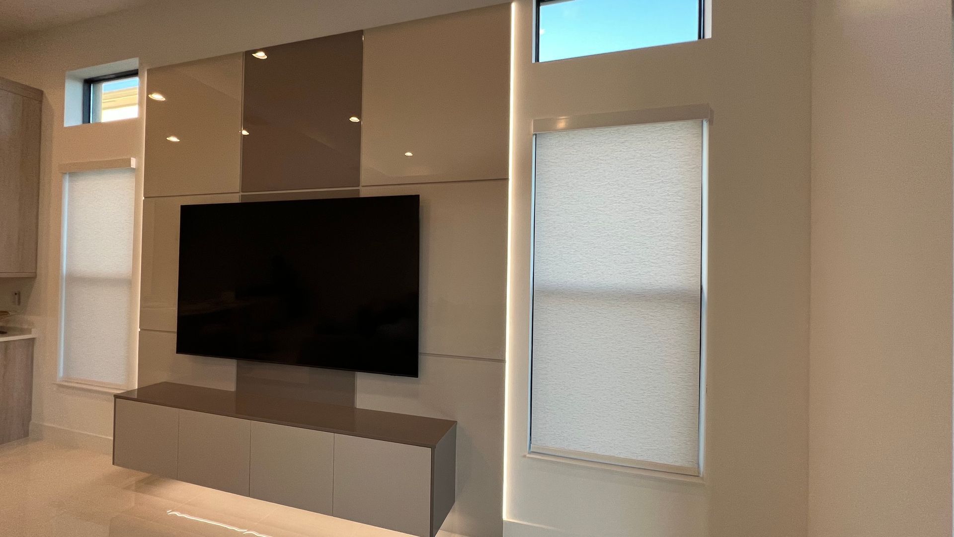 A wall-mounted TV sits above a floating console, flanked by windows with shades and accented by vertical LED strip lighting.