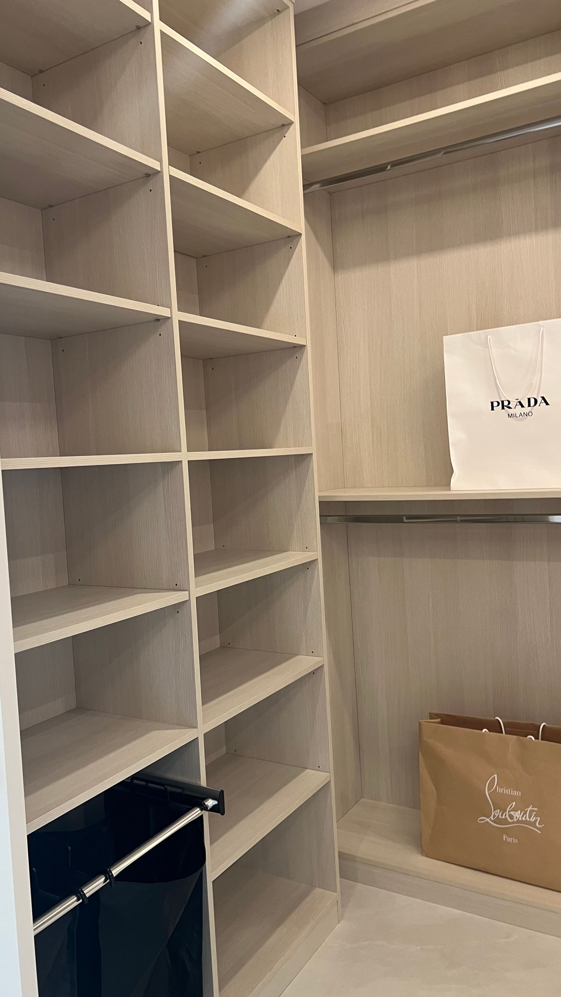 A walk-in closet with light wood shelving, a hanging rod, a white Prada shopping bag, and a brown Christian Louboutin bag.
