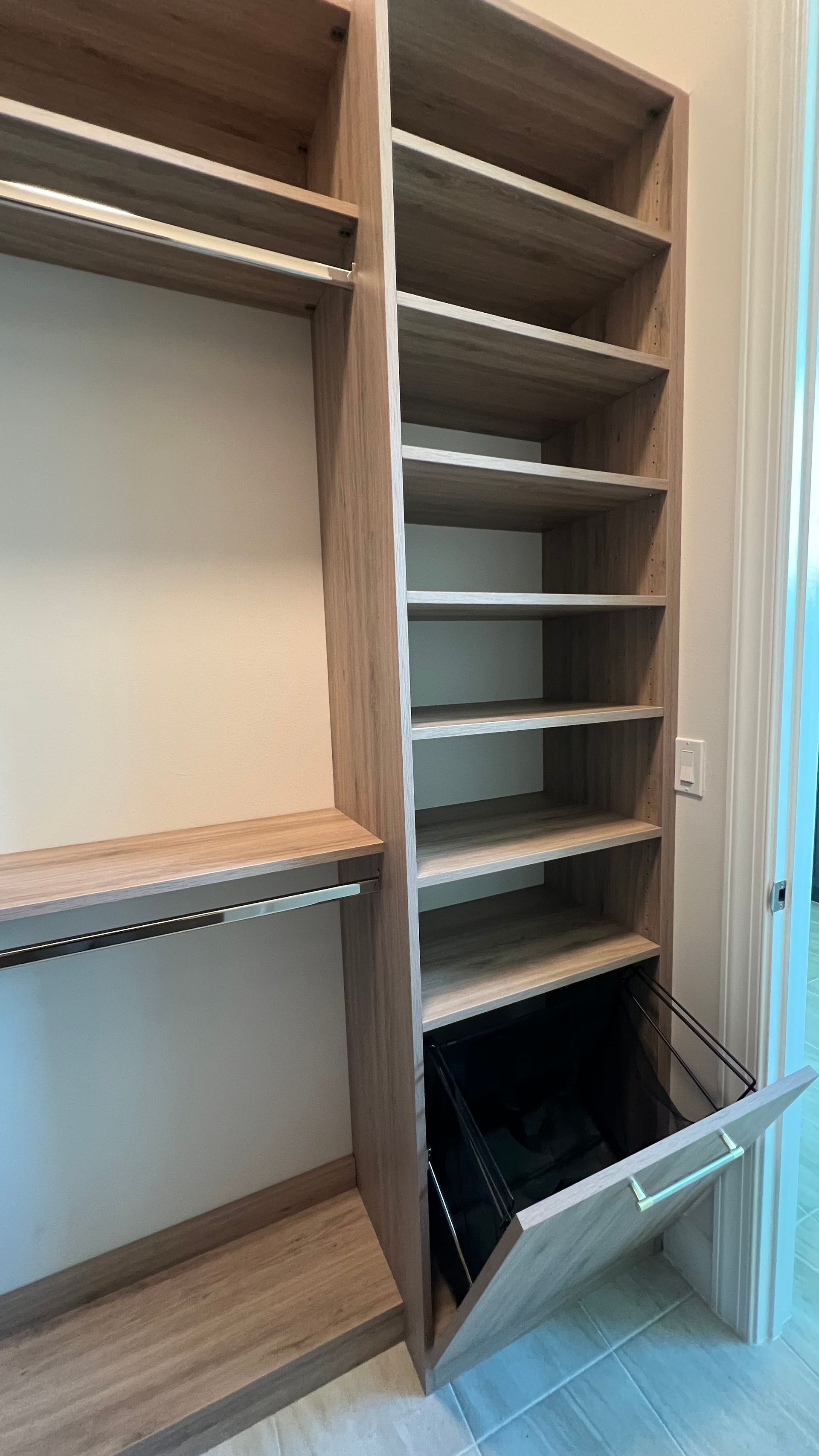 A light wood-toned closet organizer with shelves and a tilted pull-out laundry hamper at the bottom.