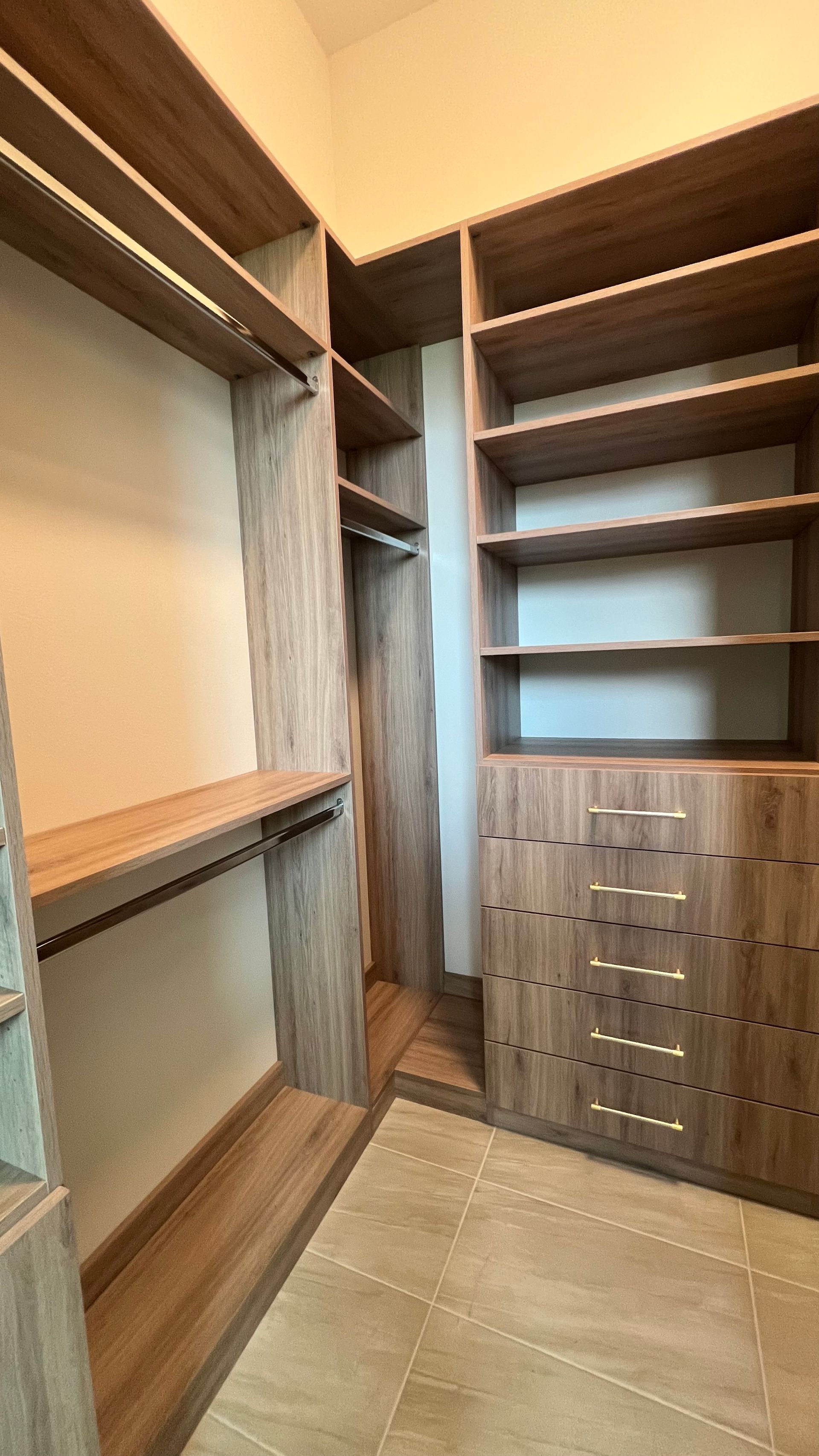 A walk-in closet with light-brown wood shelving, hanging rods, and a column of drawers on a tile floor.
