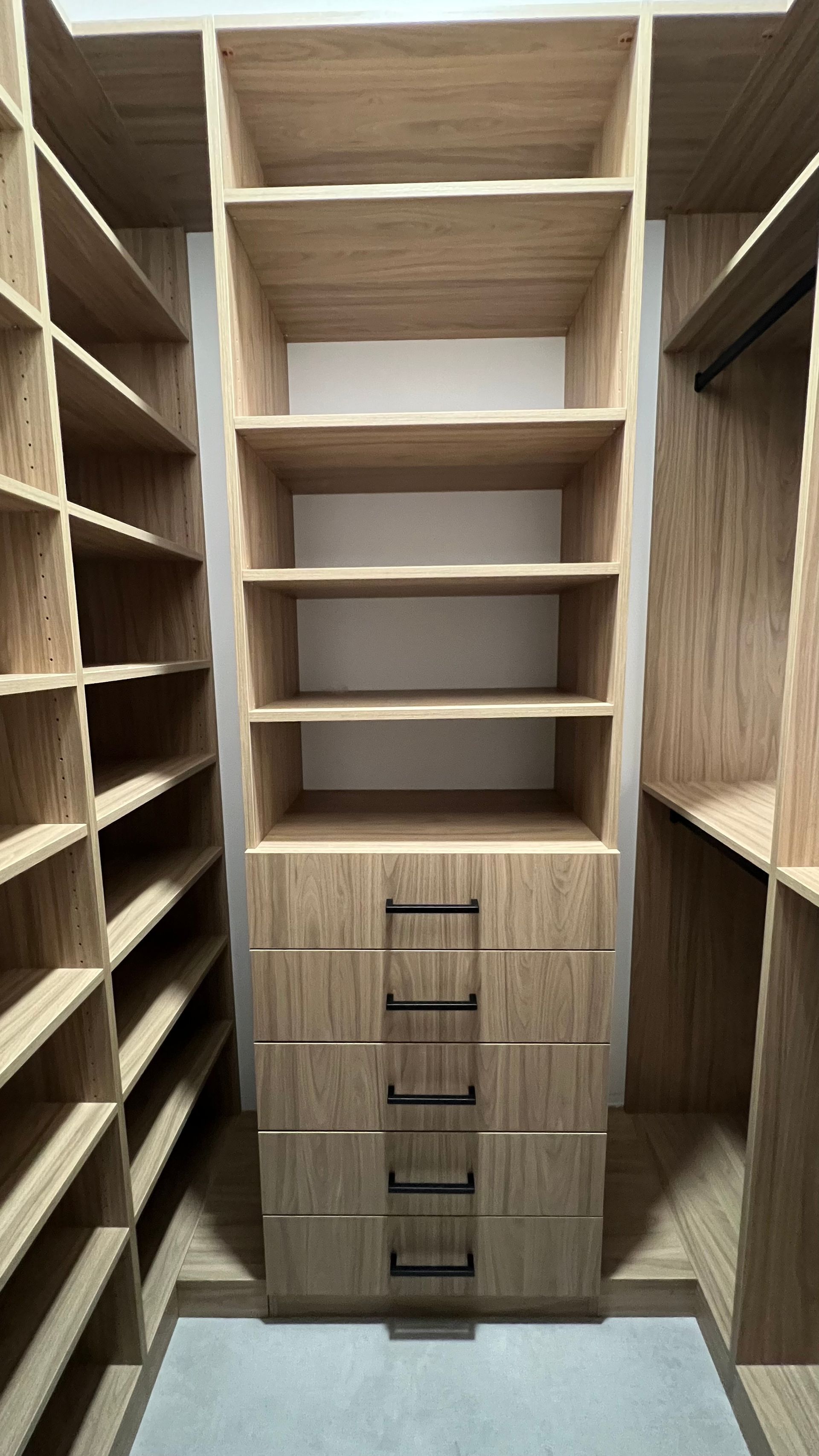 Custom light-wood walk-in closet featuring open shelving, a five-drawer stack with black handles, and hanging space.