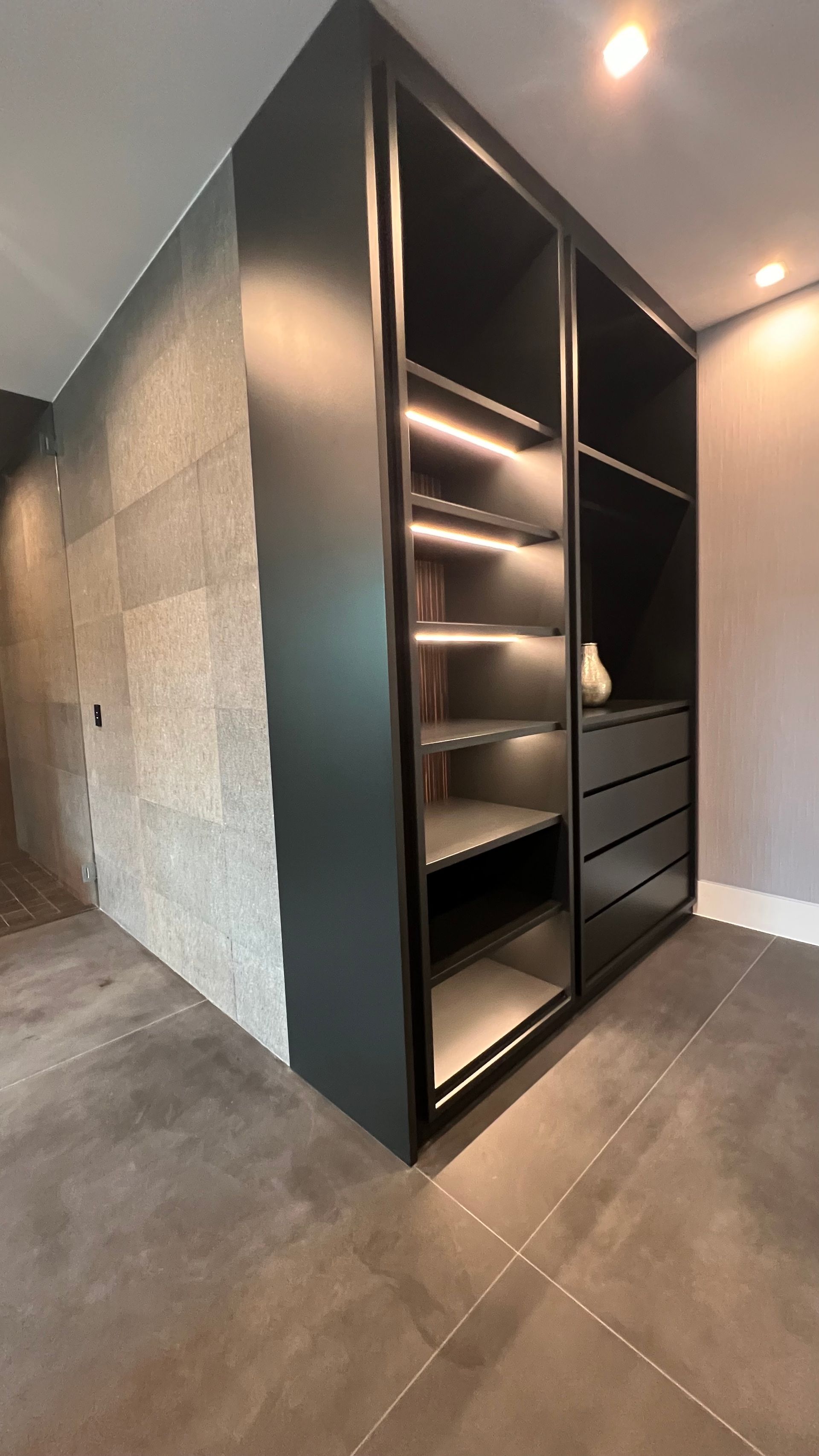 Modern dark grey wardrobe with integrated LED-lit shelves and drawers, positioned against a textured stone wall.