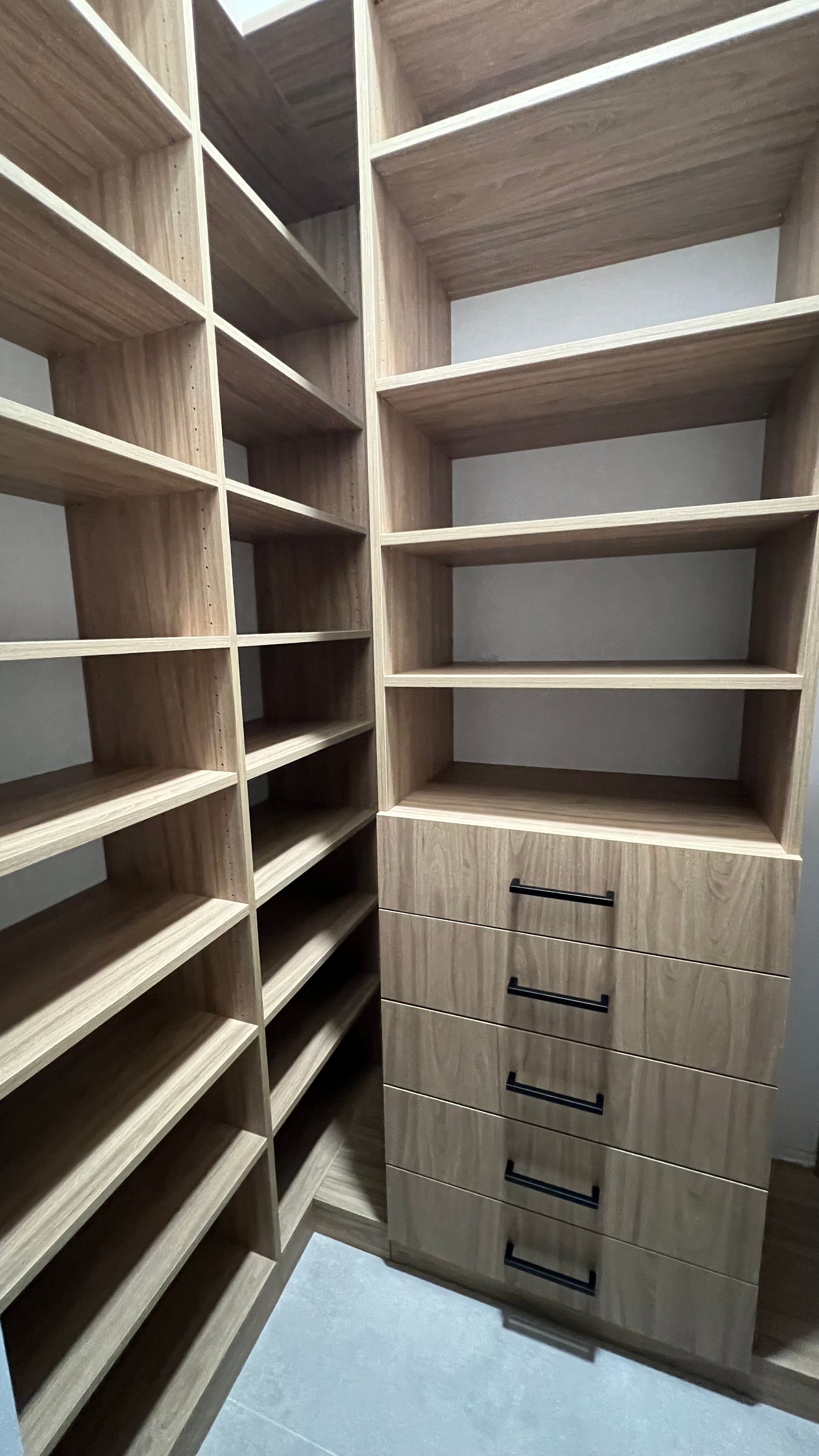 A walk-in closet with light-wood shelves and a central column of drawers featuring black horizontal handles.