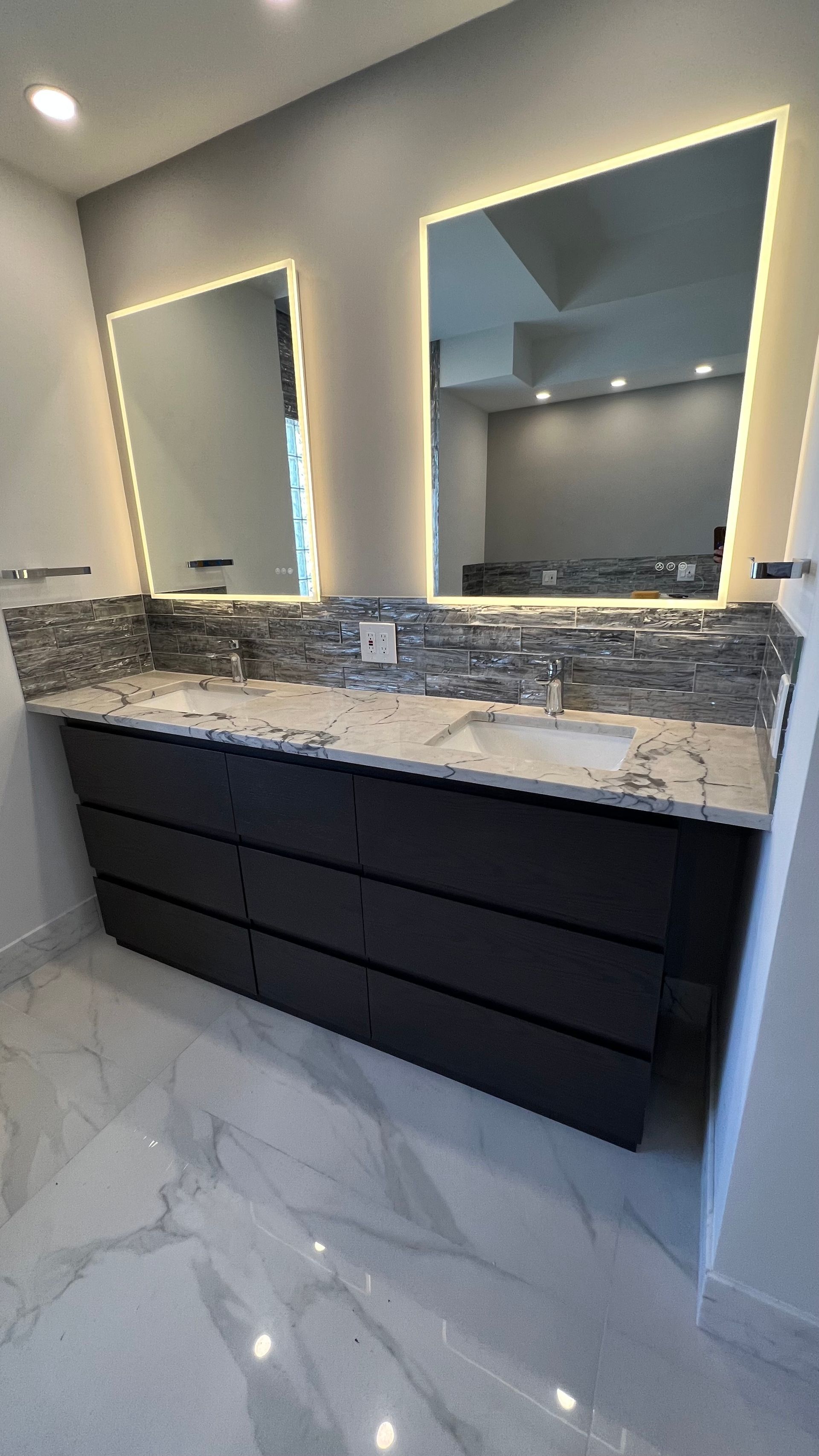 Modern double vanity with dark cabinets, marble-patterned countertop, stone backsplash, and two backlit mirrors.