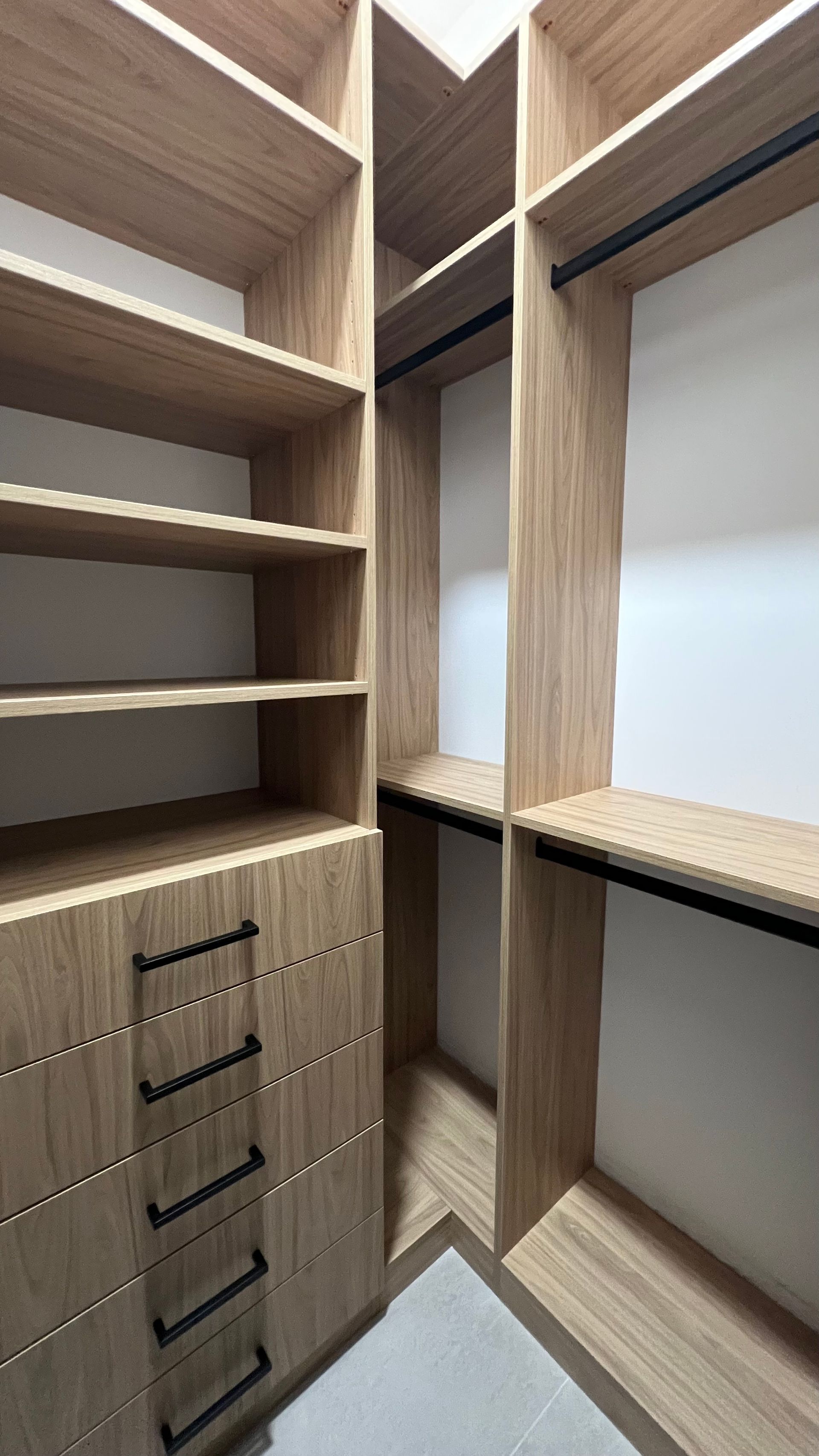 A modern walk-in closet with light wood shelving, hanging rods, and a column of five drawers with black metal handles.