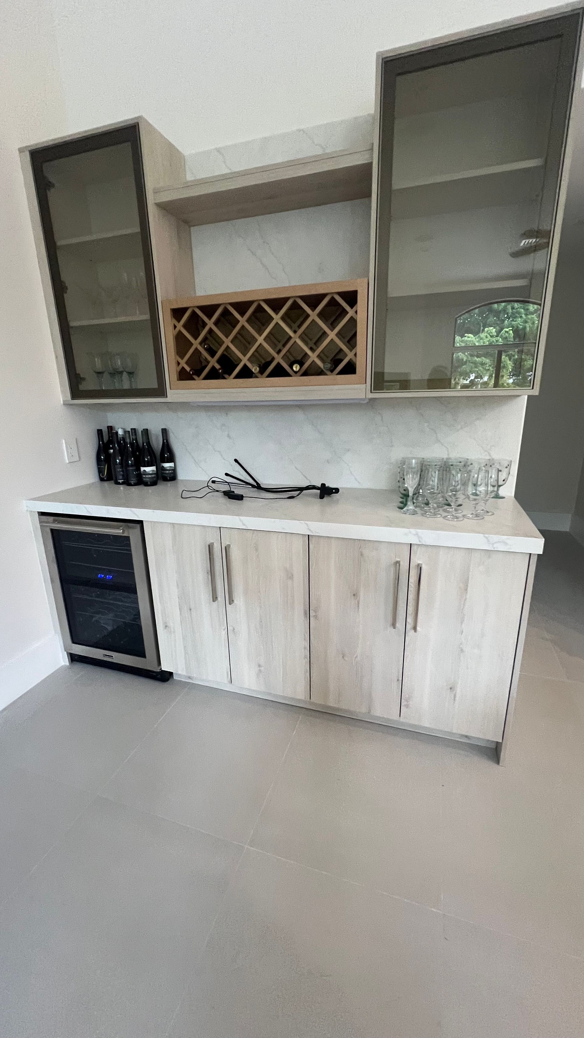 Light-colored wood home bar cabinet with glass upper cabinets, a wine rack, and a built-in wine cooler on a light floor.