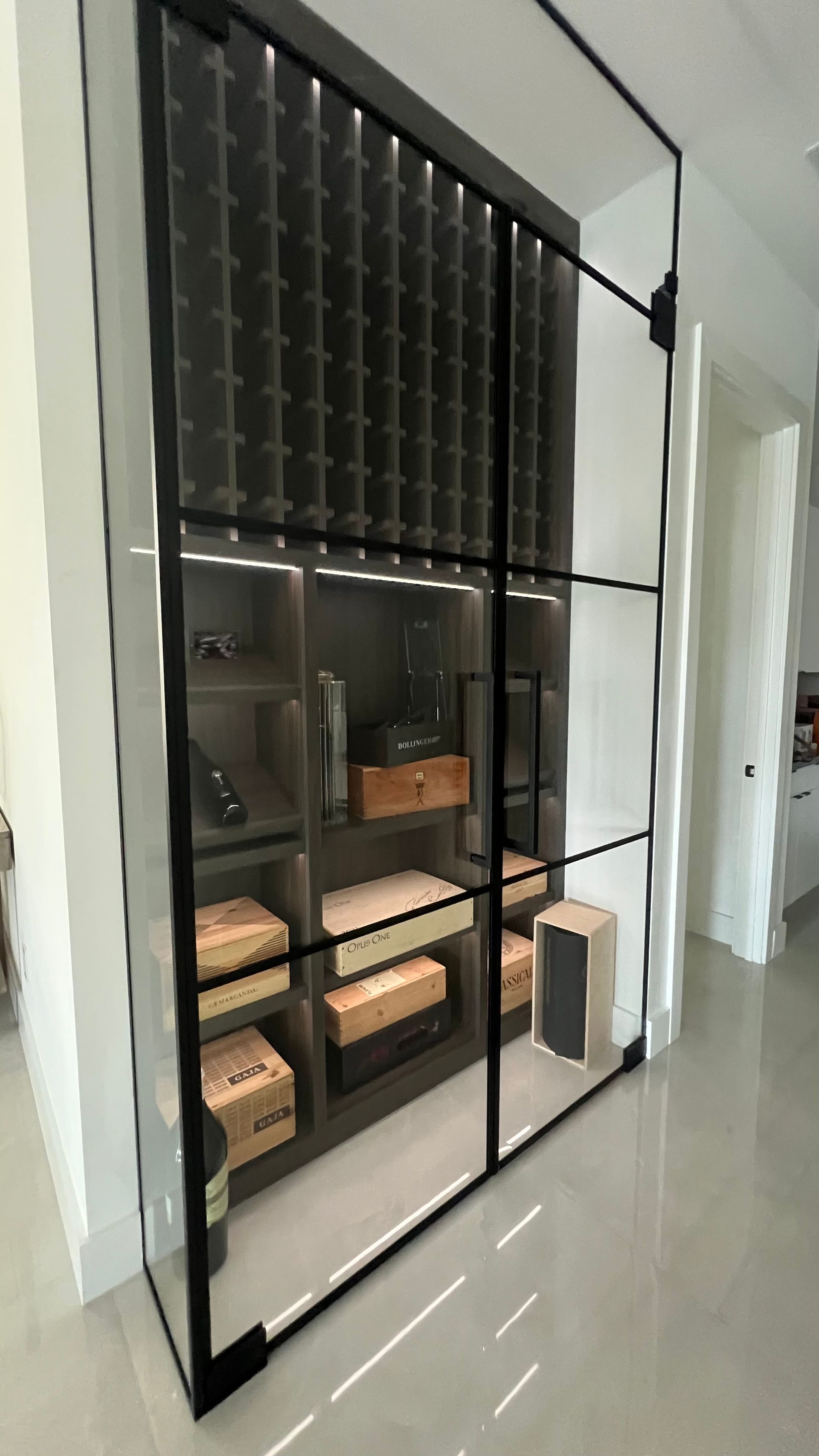 A modern, floor-to-ceiling glass wine cabinet with black metal framing and interior LED lighting in a minimalist home.