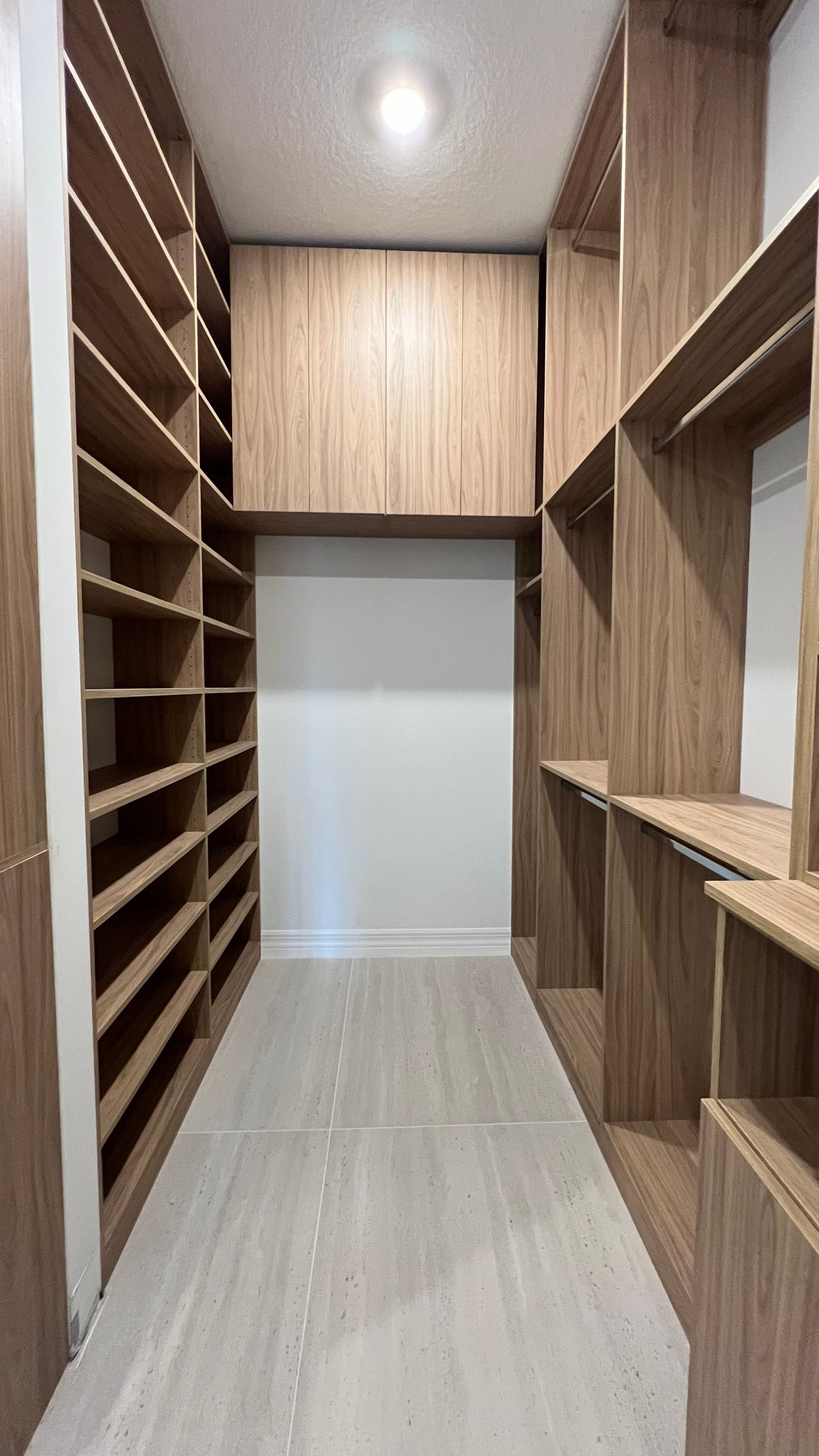 A walk-in closet featuring light-wood shelving and cabinetry, a high overhead cabinet, and light-colored tile flooring.