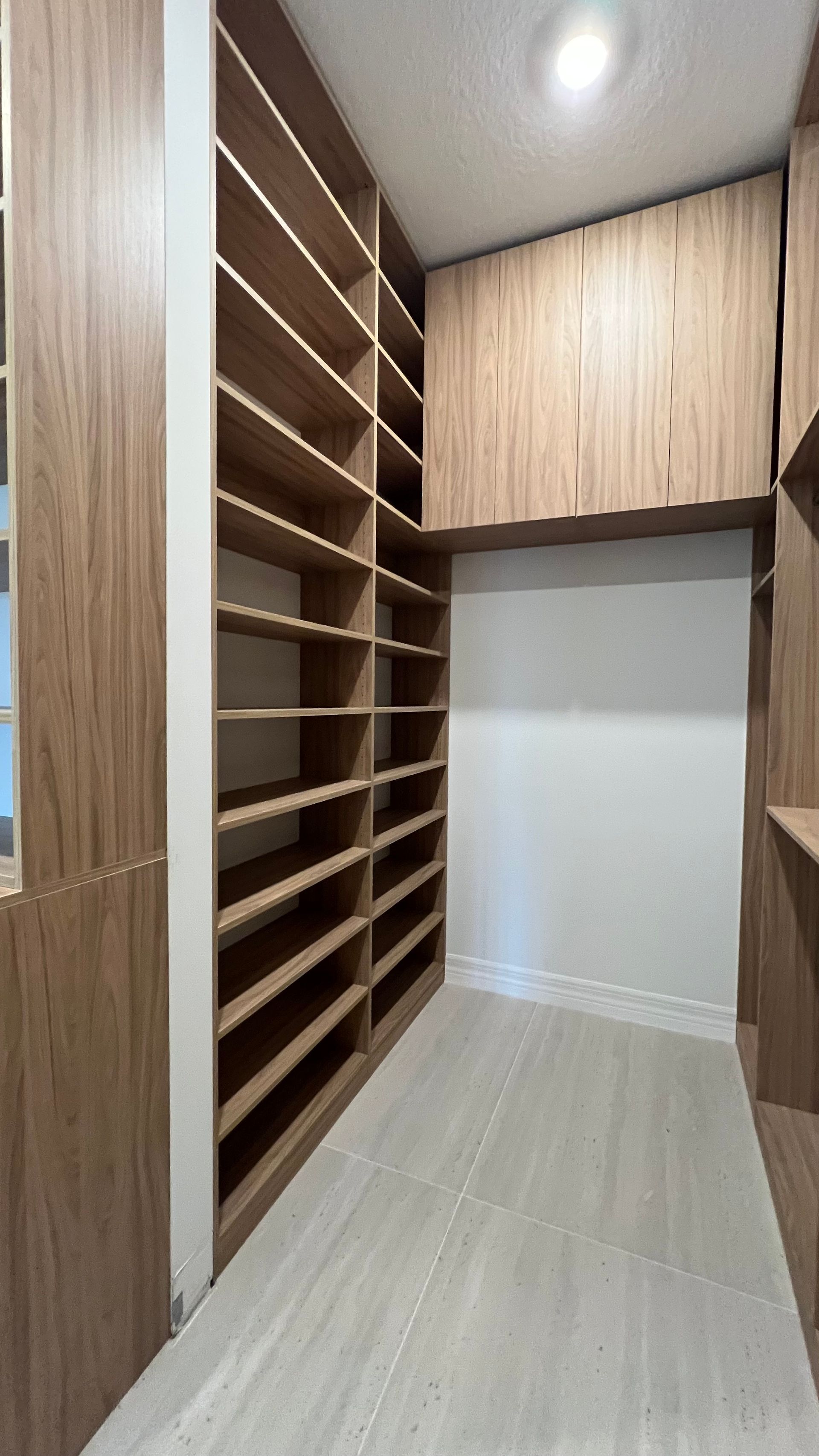 A walk-in closet featuring floor-to-ceiling wooden shelving units, a high cabinet, and light-colored tiled flooring.