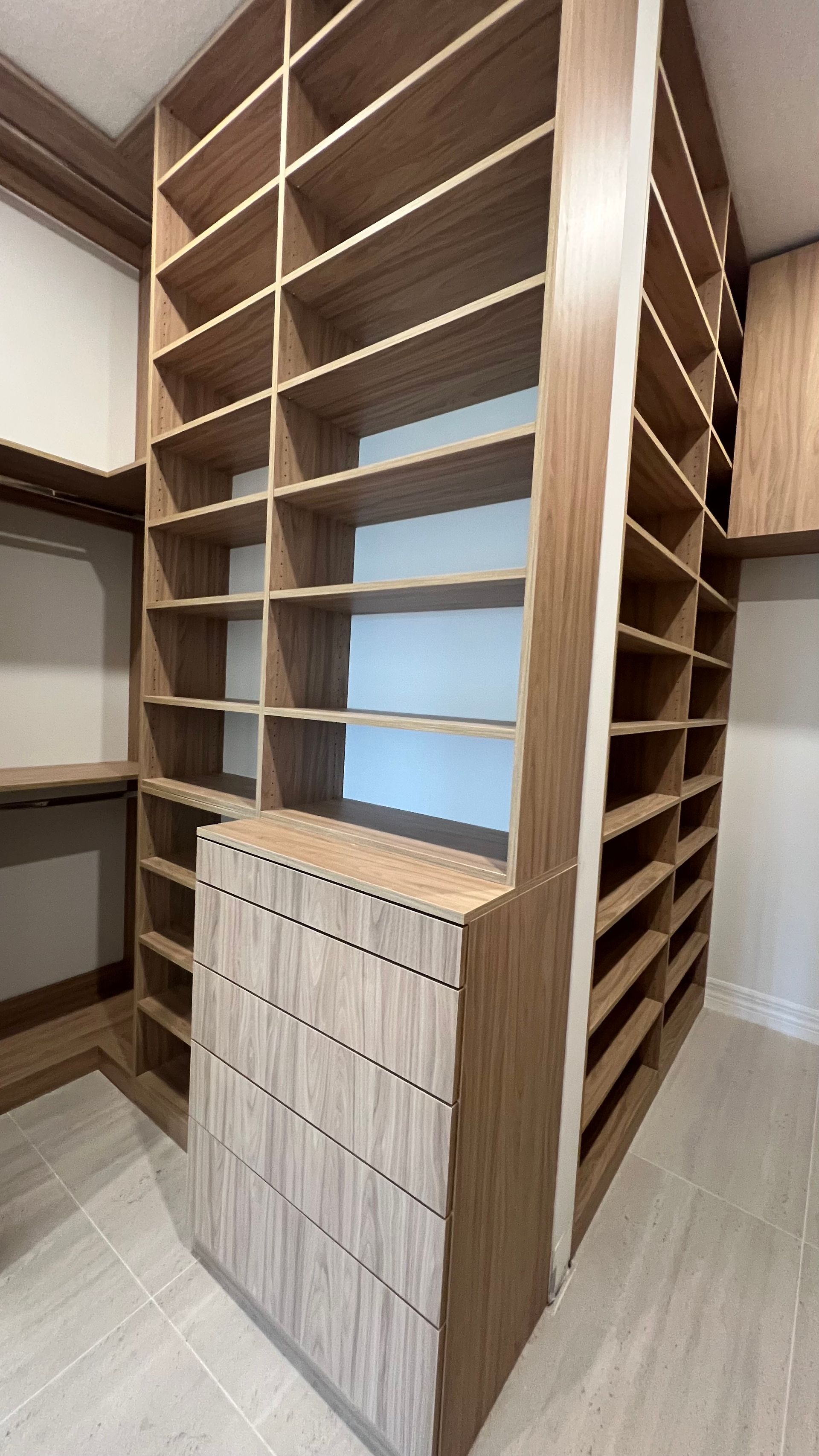 Modern, light-wood walk-in closet featuring tall shelving units and a chest of five drawers on a tiled floor.
