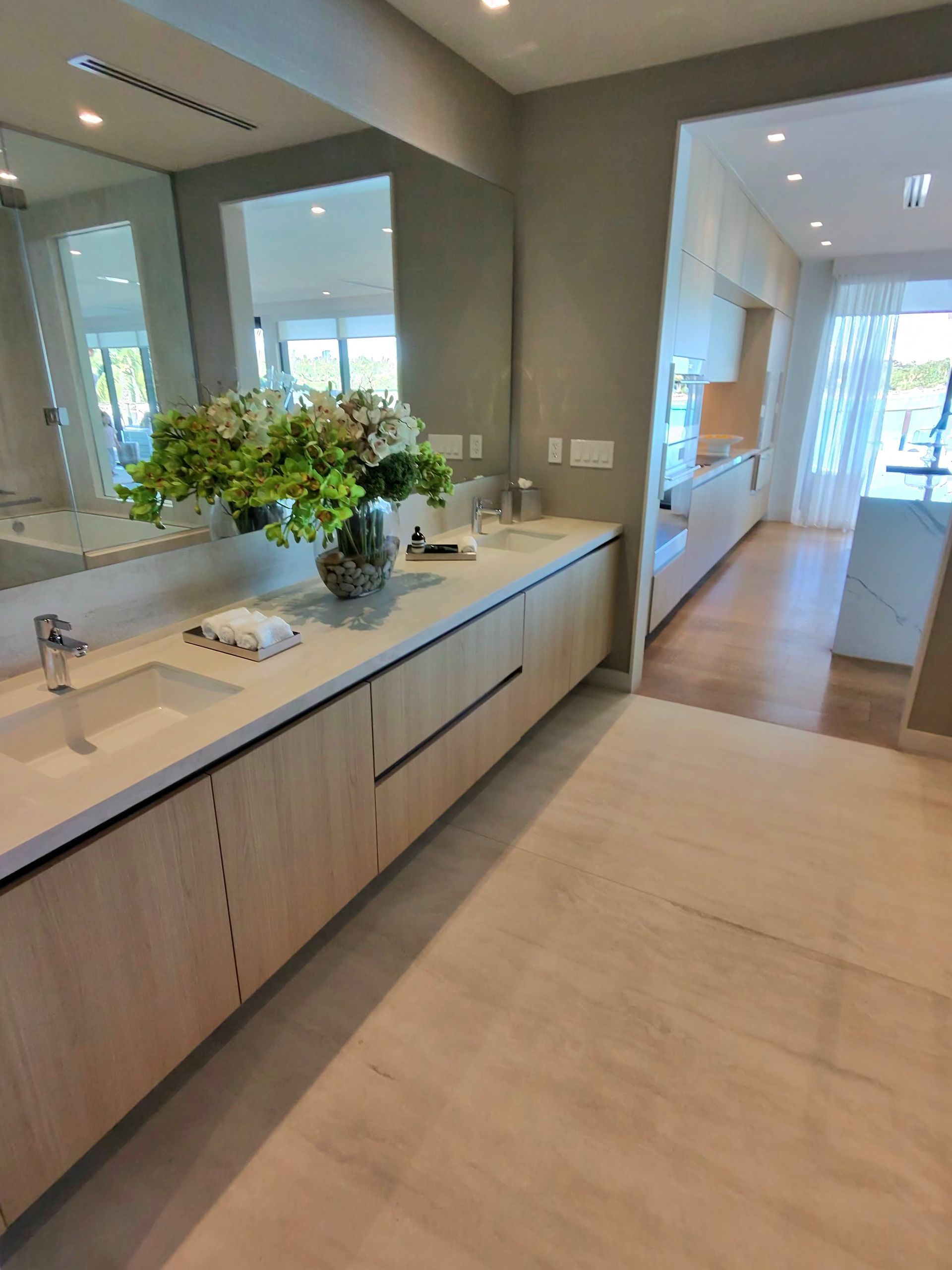 A modern bathroom vanity with light wood cabinetry, a large mirror, a vase of green flowers, and a view into a hallway.
