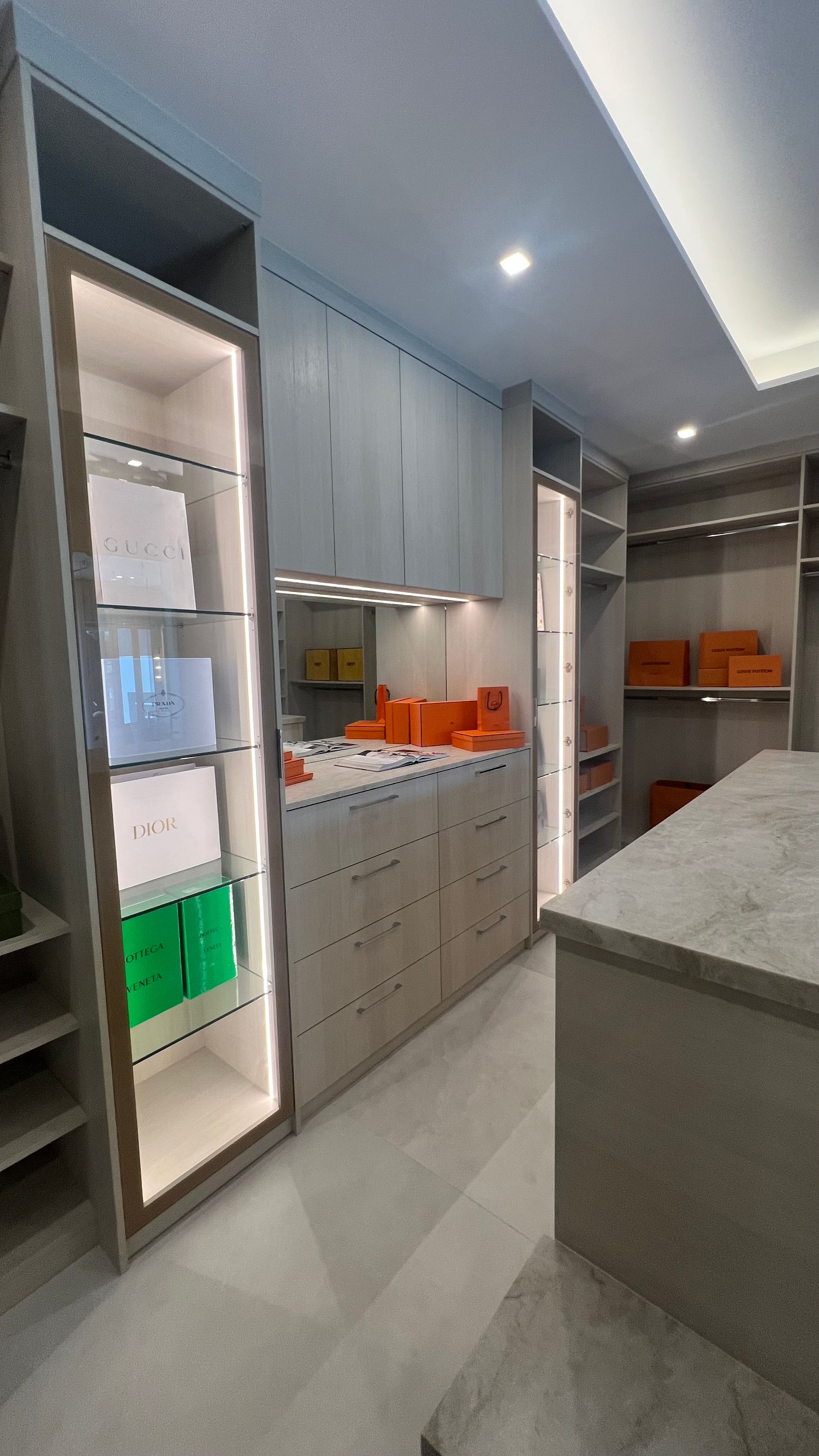 A modern walk-in closet featuring light gray cabinetry, LED-lit glass shelving, and a marble-topped center island.