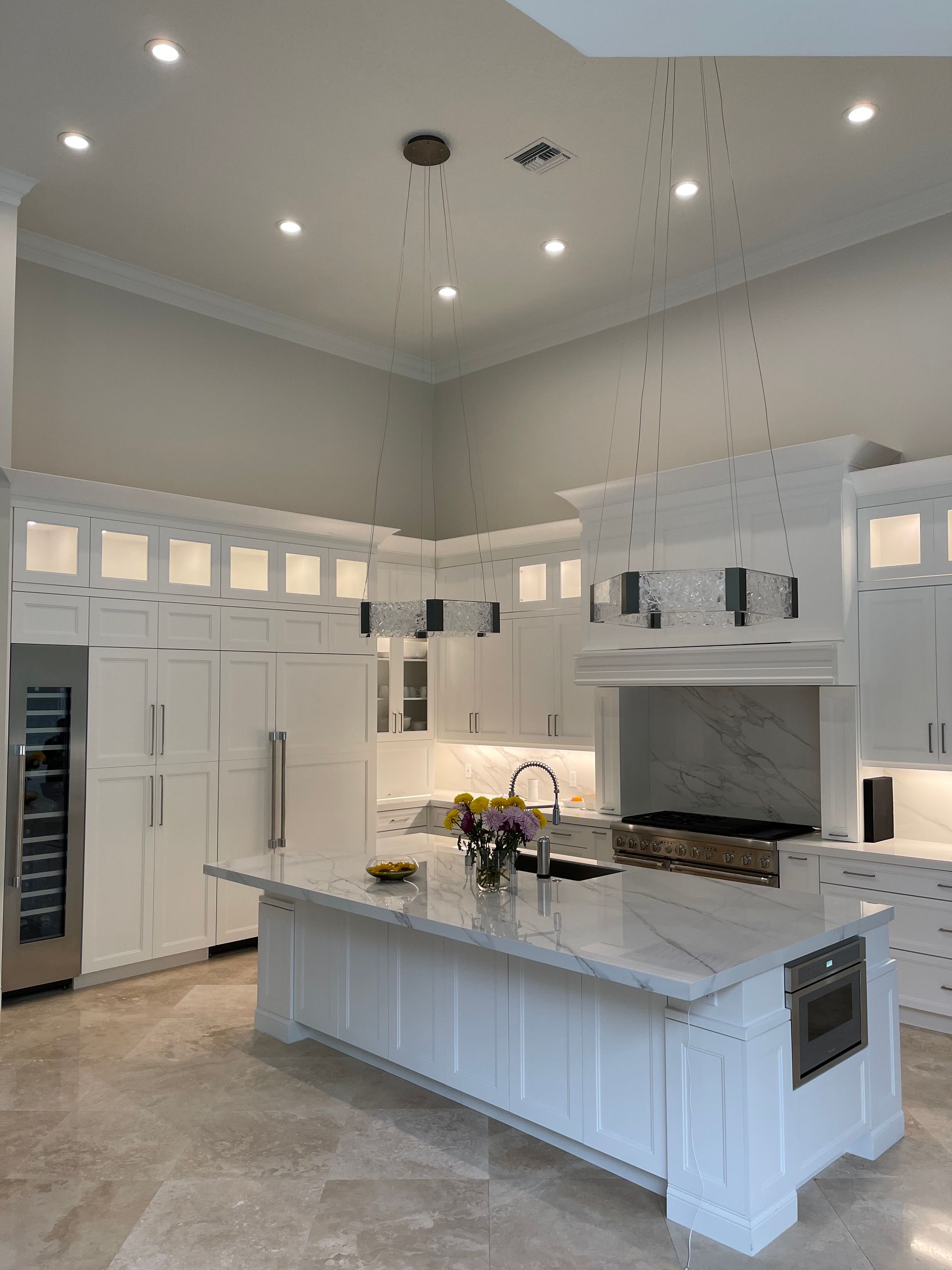 A bright, modern kitchen featuring white cabinets, a large marble island with seating, pendant lights, and tile floors.