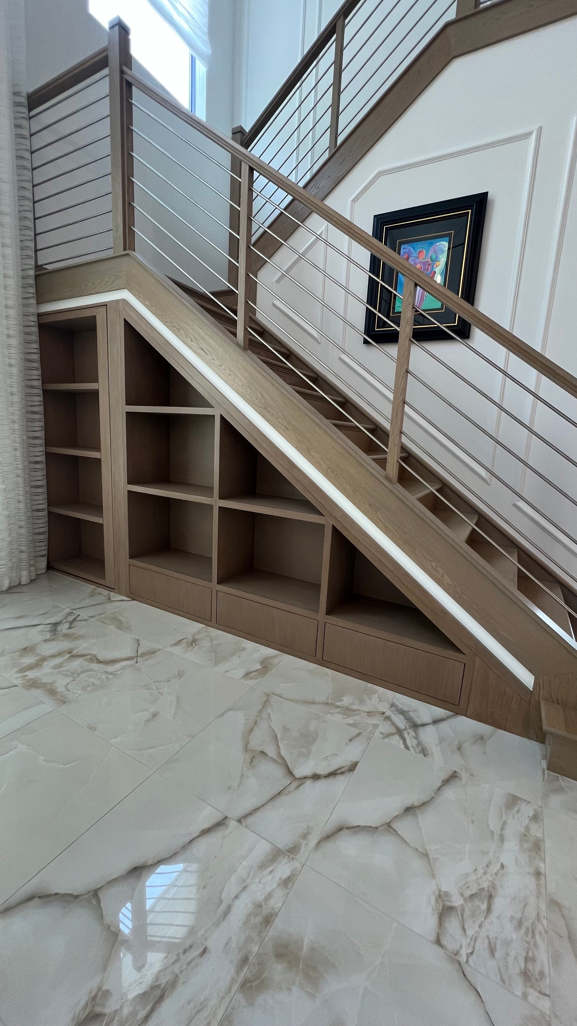 A wooden staircase with cable railings features built-in, open shelving underneath in a home with large marble floor tiles.