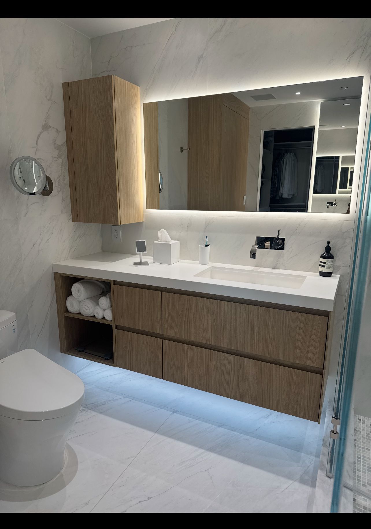 Modern bathroom featuring a wood vanity with under-cabinet lighting, white marble walls, and a large illuminated mirror.