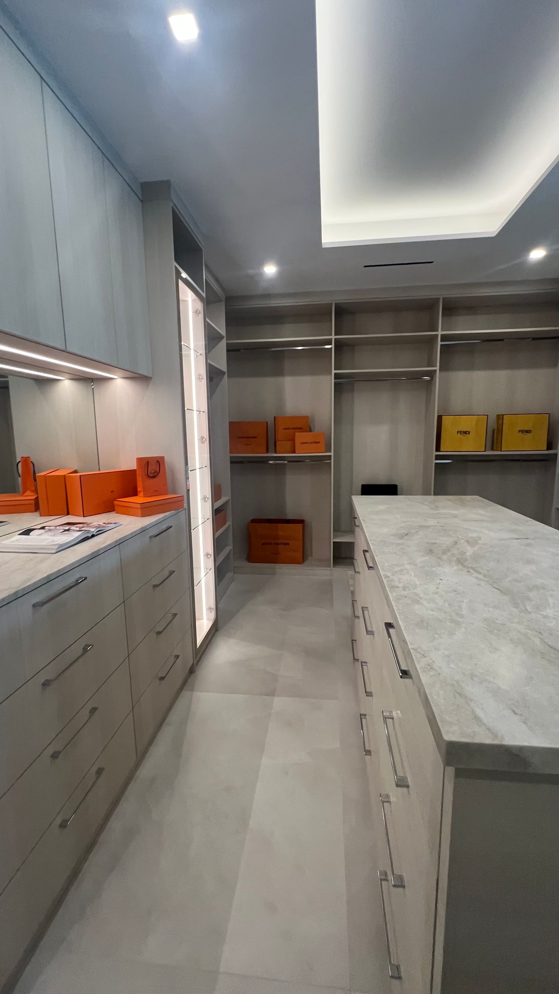 A modern, minimalist walk-in closet with light wood cabinetry, white marble countertops, and orange storage boxes.