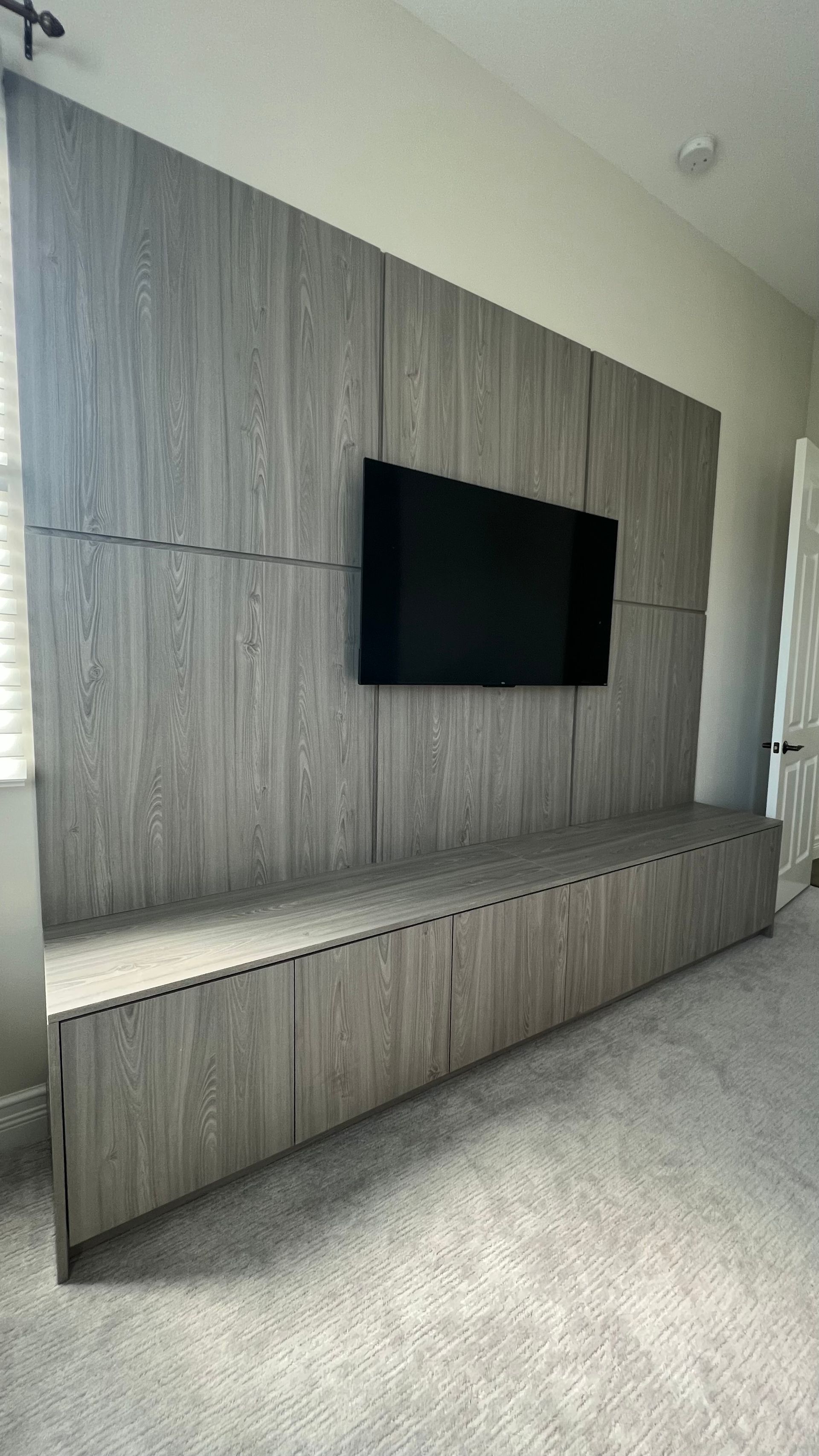 Wall-mounted TV above a long, gray wood-grain console unit in a room with light carpeting.