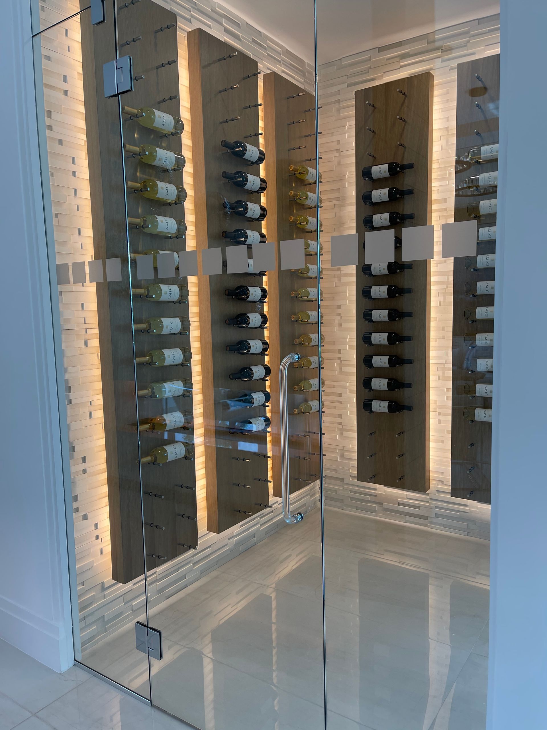 A modern glass-enclosed wine cellar with bottles displayed on vertical wood panels illuminated by soft backlighting.