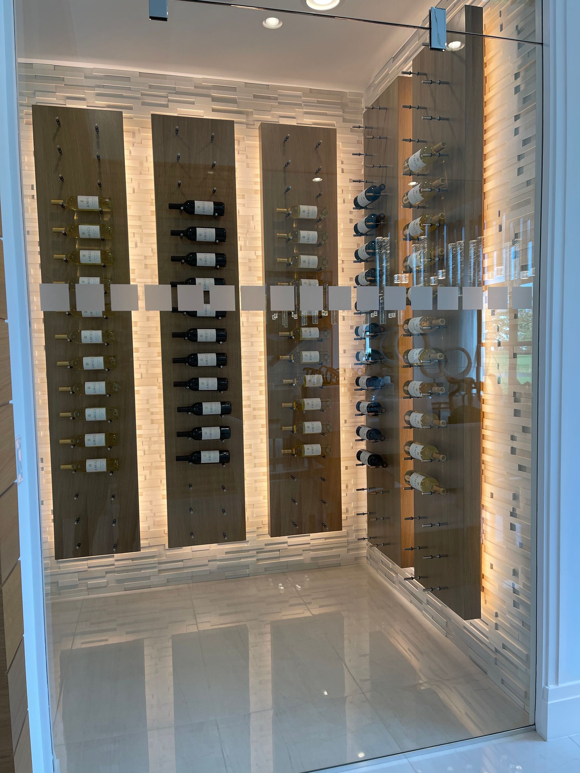 A modern glass-enclosed wine cellar with illuminated vertical wood panels holding horizontal wine bottles.
