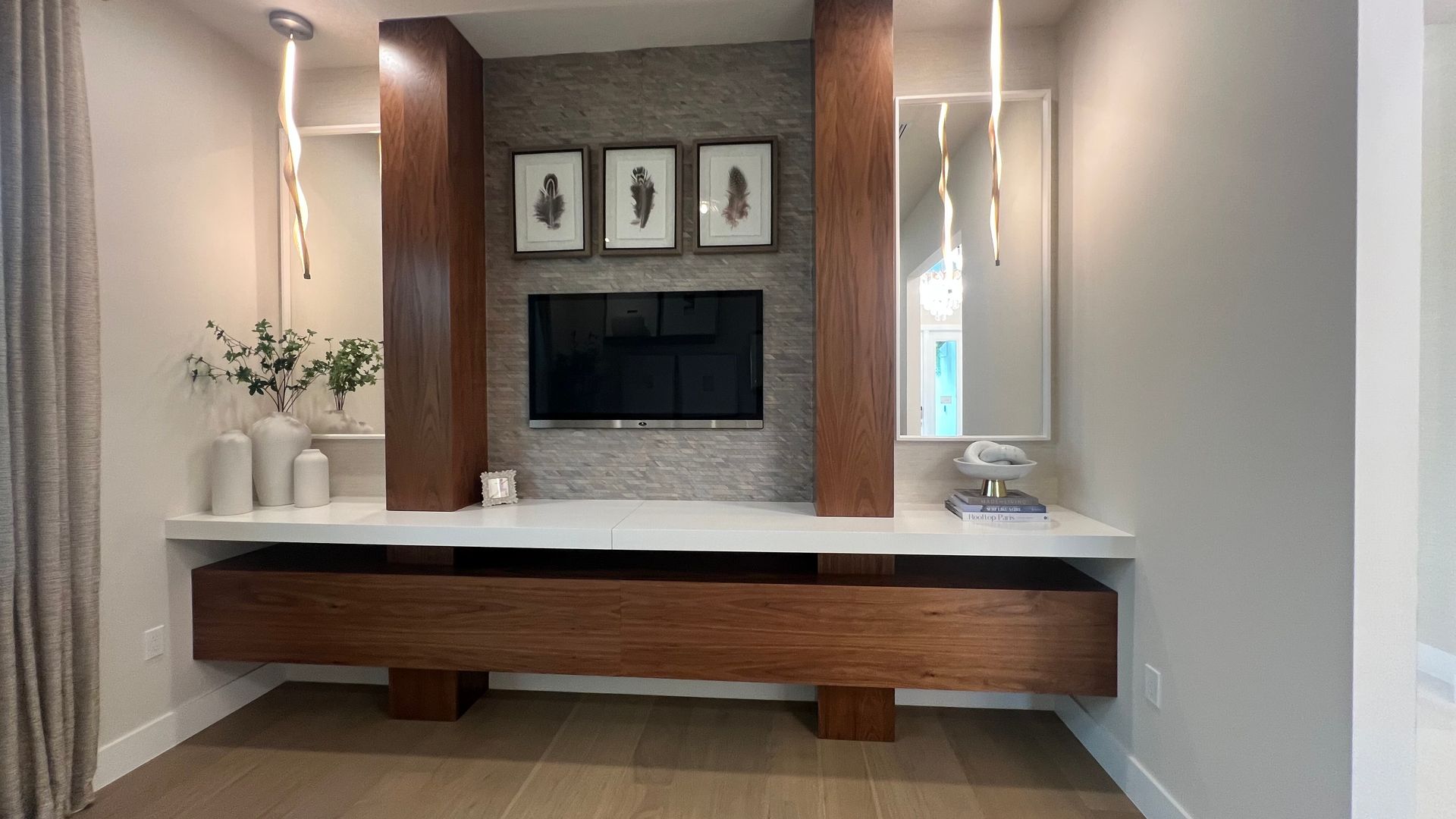 A modern living room wall featuring a TV mounted on stone, flanked by wood columns, a white shelf, and hanging lights.
