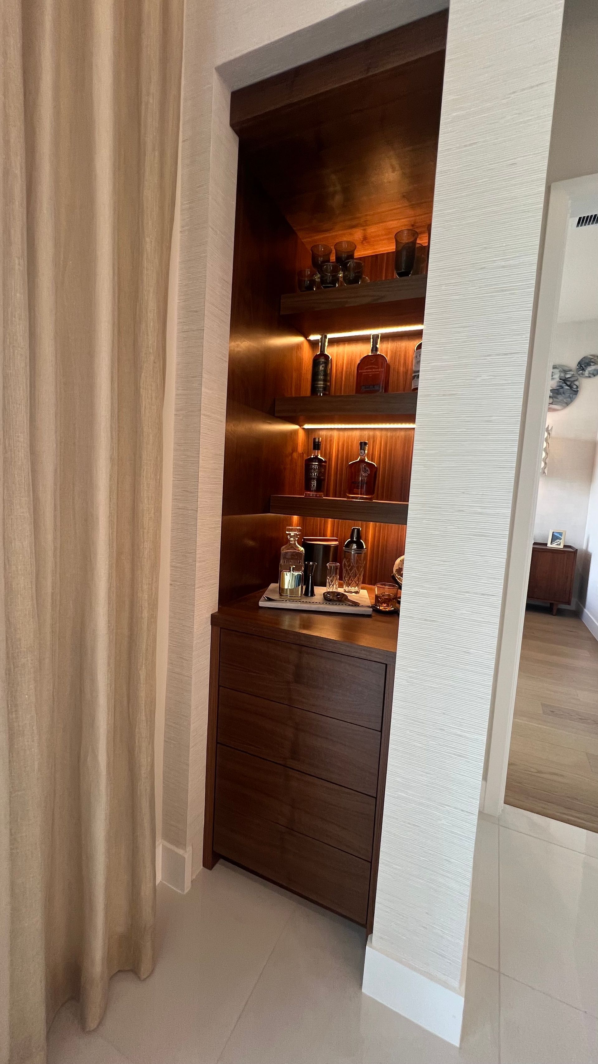 A built-in, dark wood bar cabinet with illuminated shelving holding liquor bottles, set into an alcove.