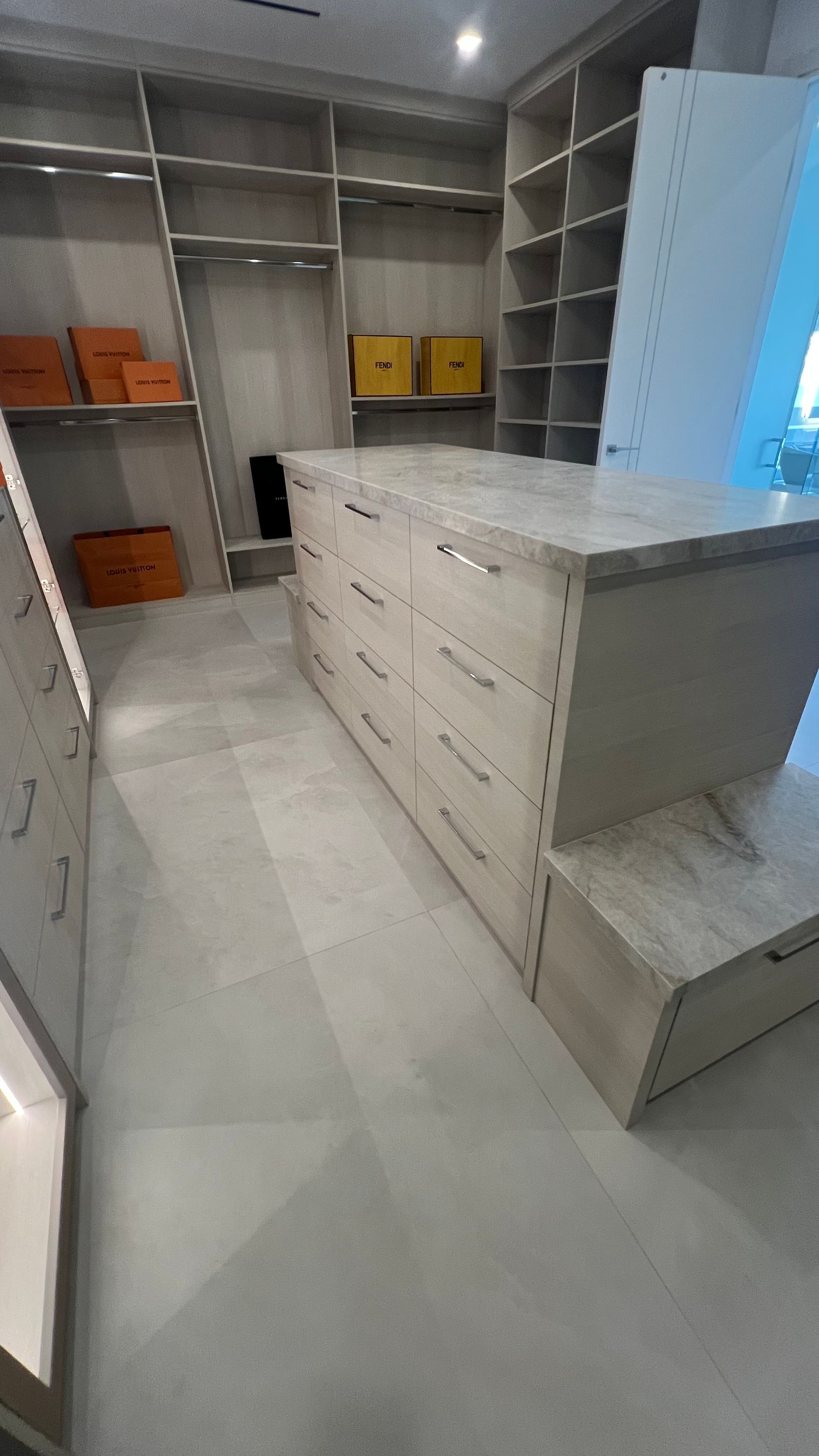 A walk-in closet featuring light-toned custom shelving, a marble-topped center island with drawers, and gray tiled flooring.