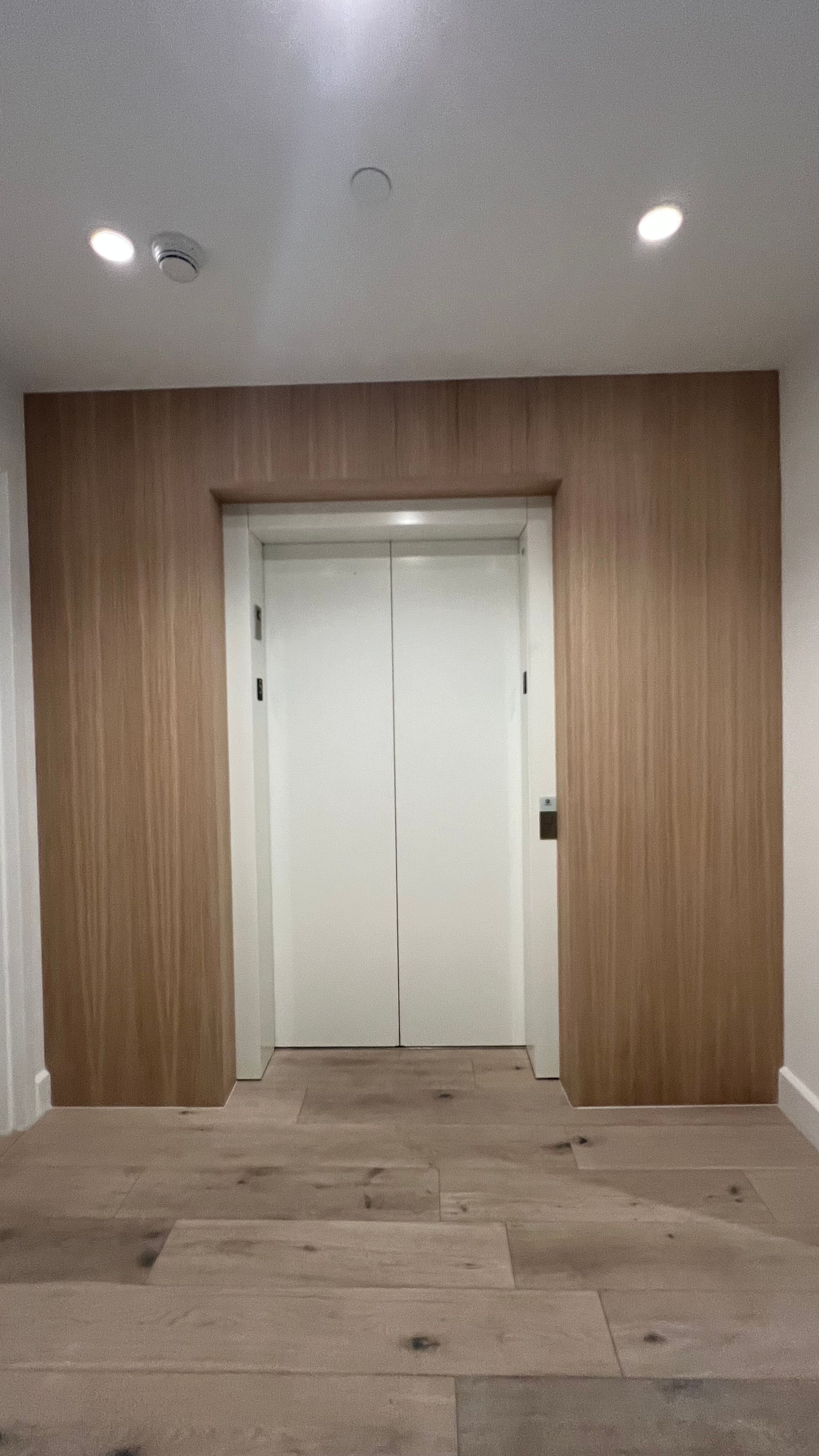 White elevator doors framed by light wood paneling in a hallway with wooden floors and two recessed ceiling lights.