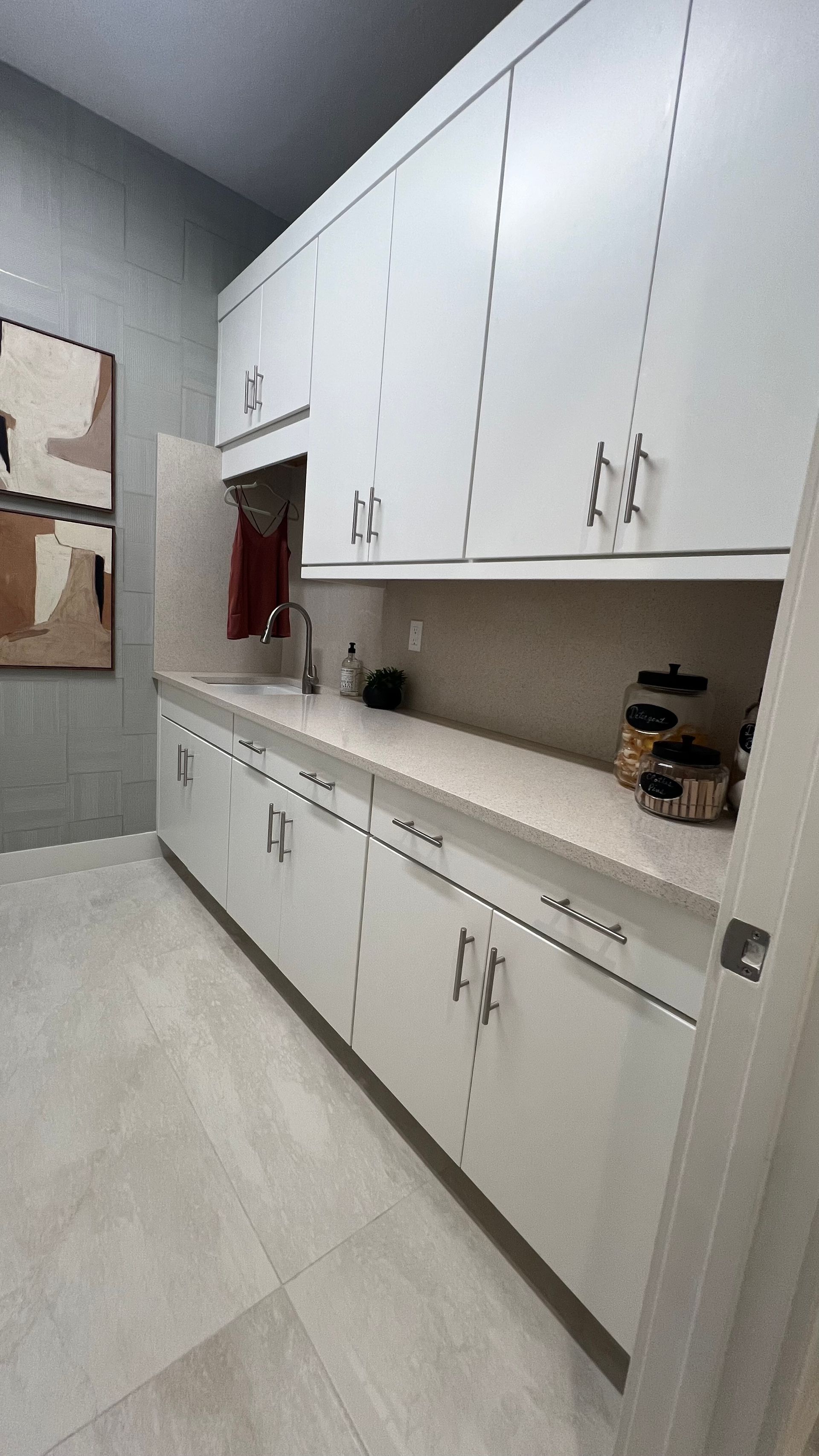 A laundry room with white upper and lower cabinets, a light countertop, a small sink, and grey-toned tile flooring.