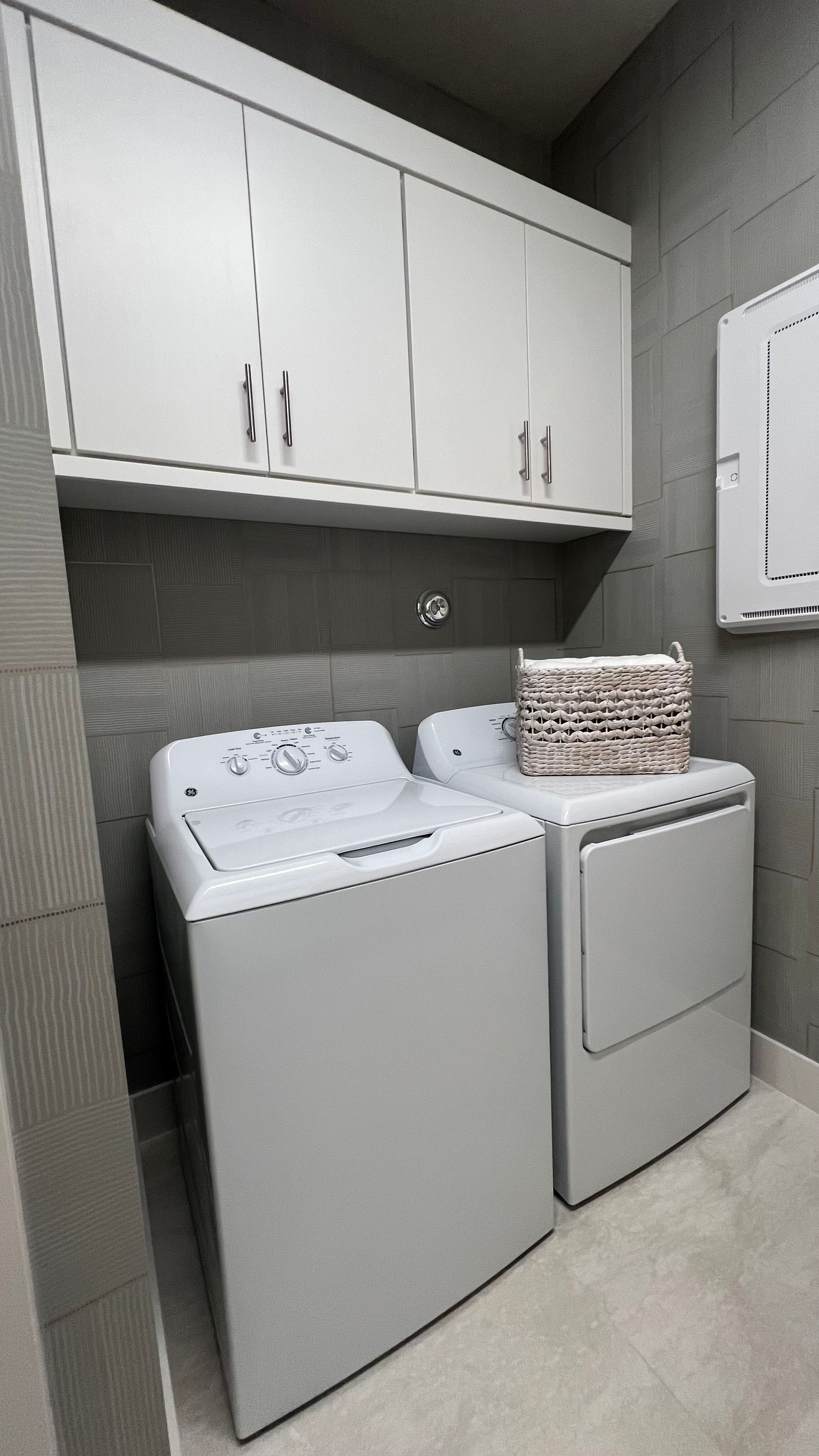 A laundry room with a white top-loading washer and dryer, a woven basket on top, and white cabinets against gray walls.
