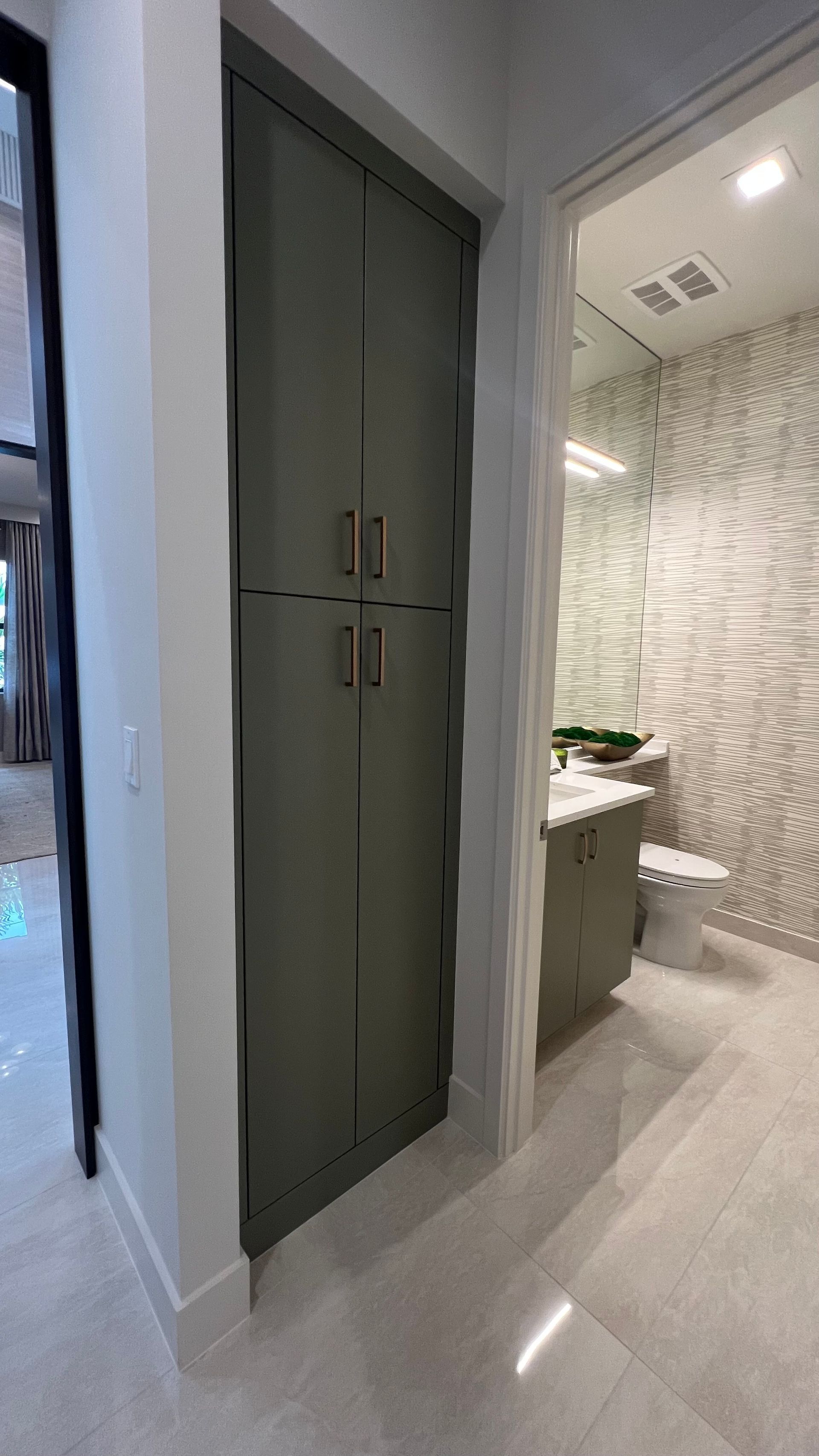 A tall, olive-green linen cabinet stands next to the entrance of a modern bathroom with a patterned wall and vanity.