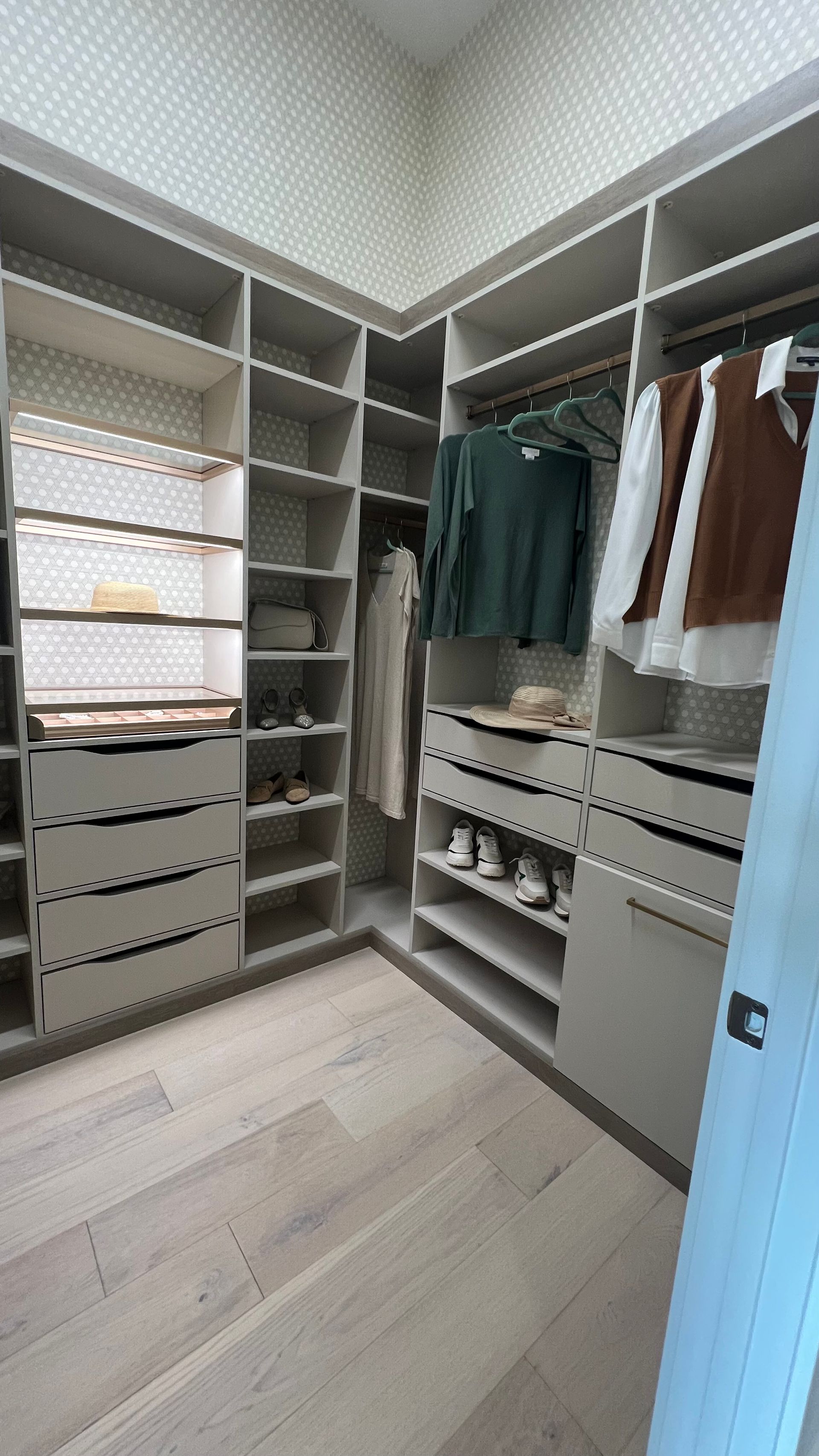 A walk-in closet featuring light gray custom shelving, a built-in shoe display with lighting, drawers, and hanging clothes.