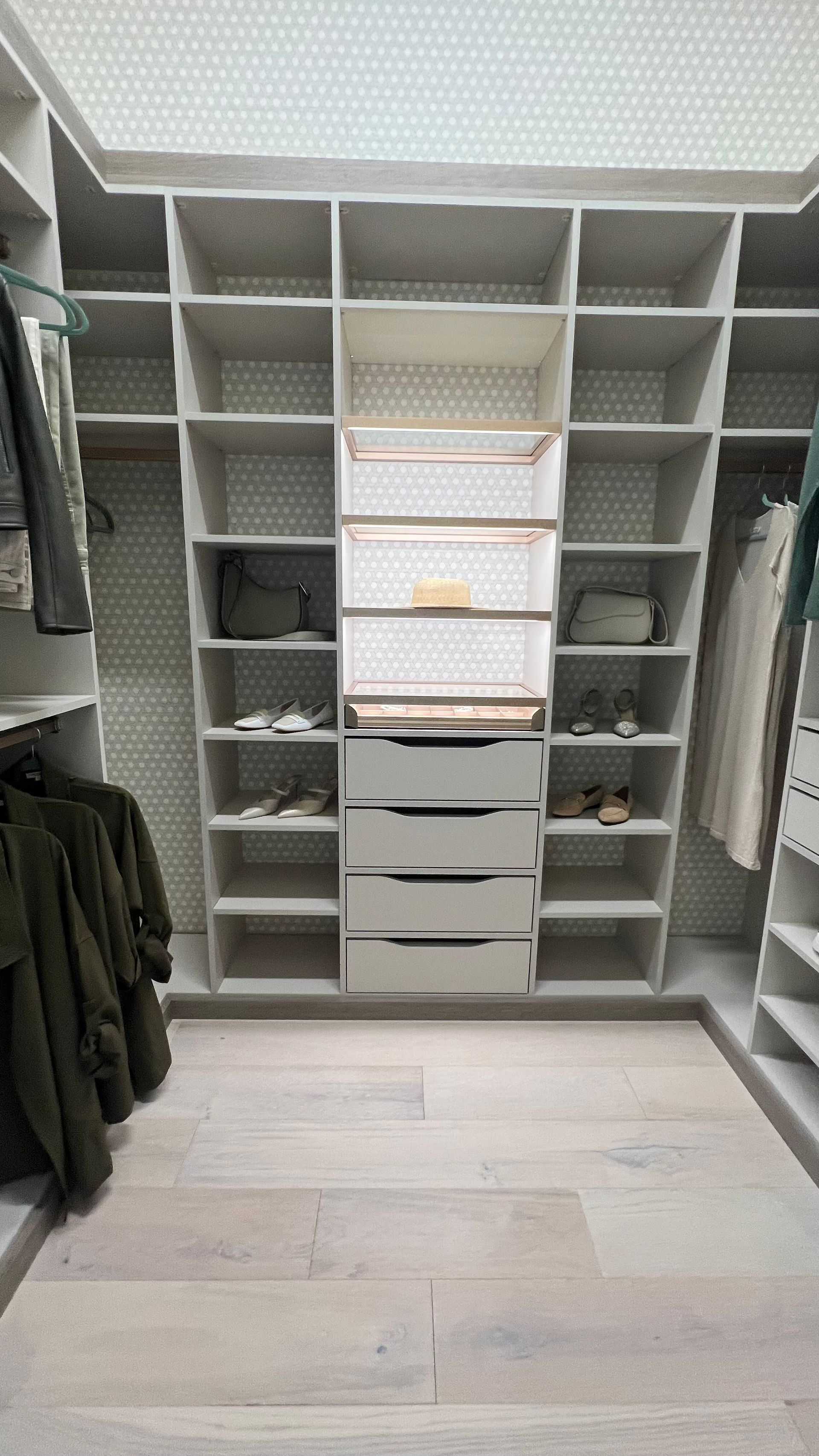 A modern, organized walk-in closet with light wood floors, white shelving, hanging rods, and a center dresser unit.