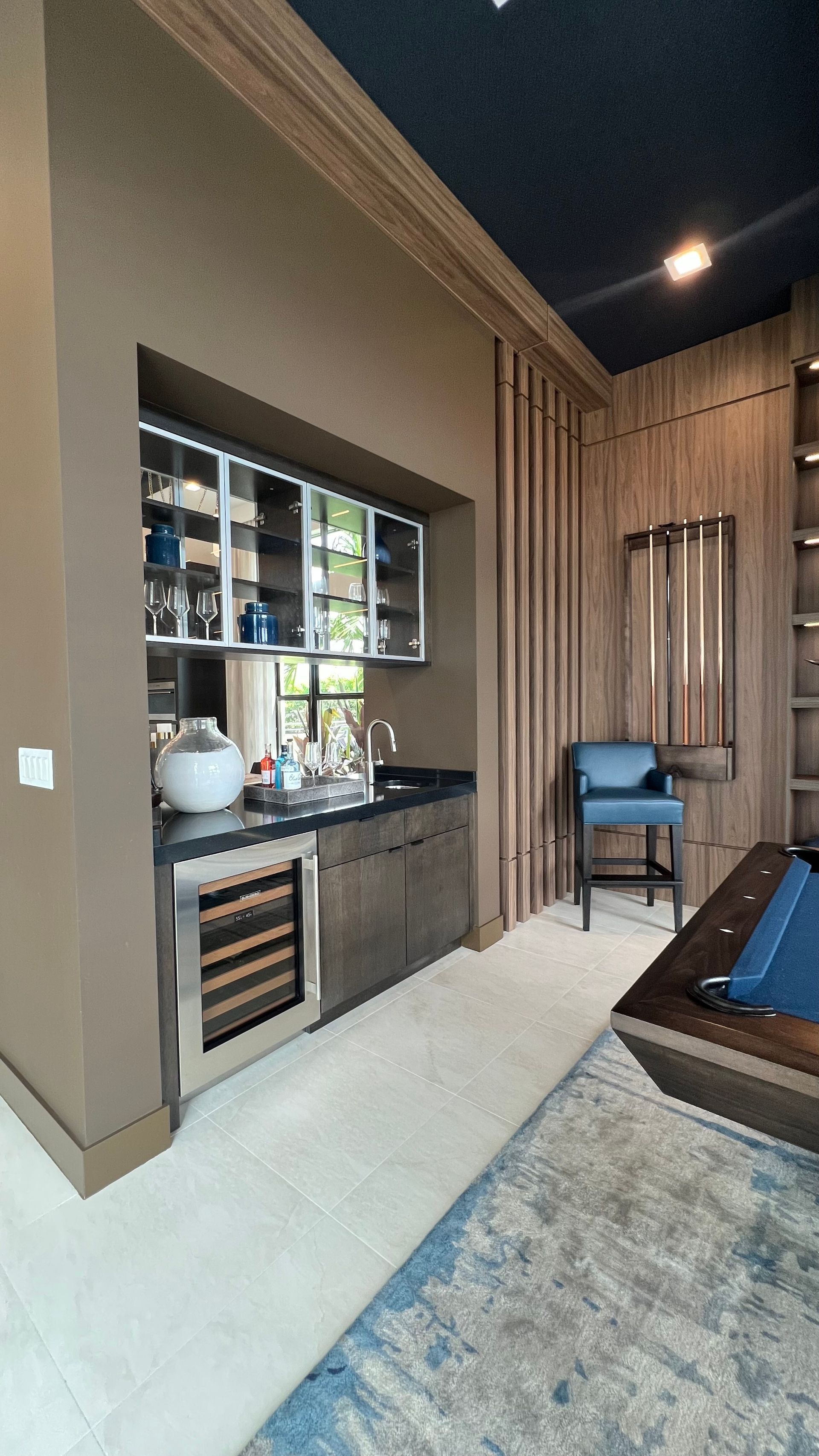 A modern wet bar area with wood-paneled walls, a wine cooler, glass-front cabinets, and a blue stool on a tile floor.