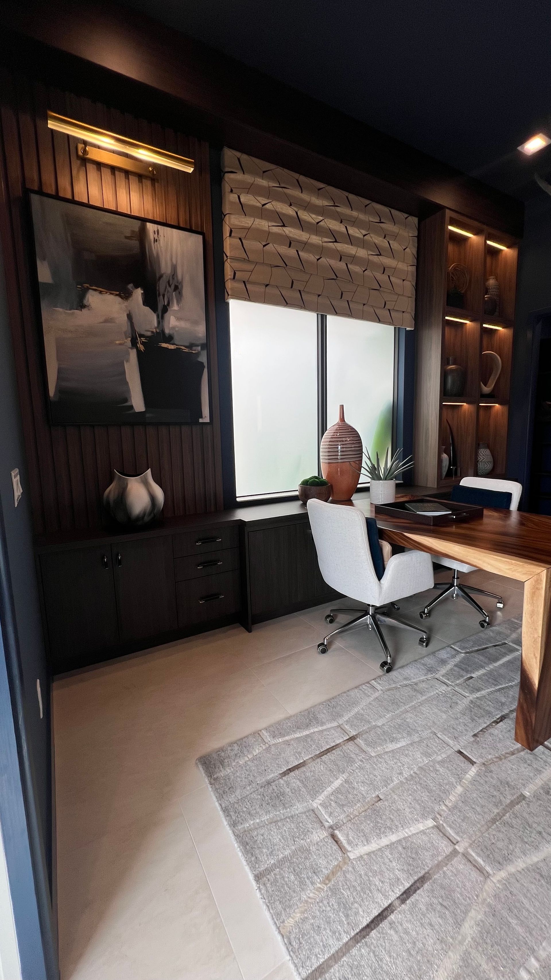 A modern office featuring a live-edge wood desk, white swivel chair, dark cabinetry, wall art, and patterned window shade.