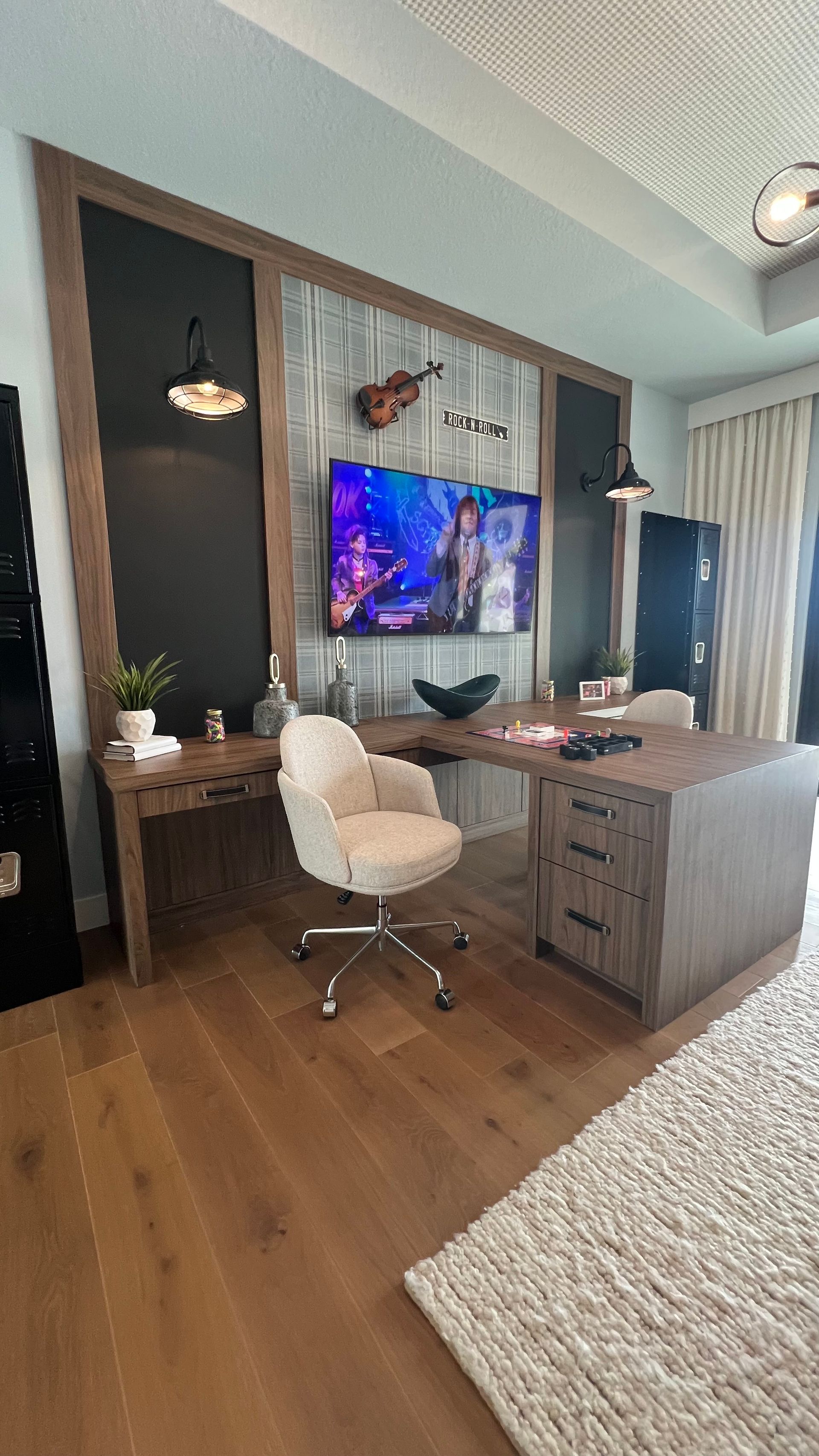 A modern home office with a wood-grain L-shaped desk, cream-colored swivel chair, and a wall-mounted TV over a rustic panel.