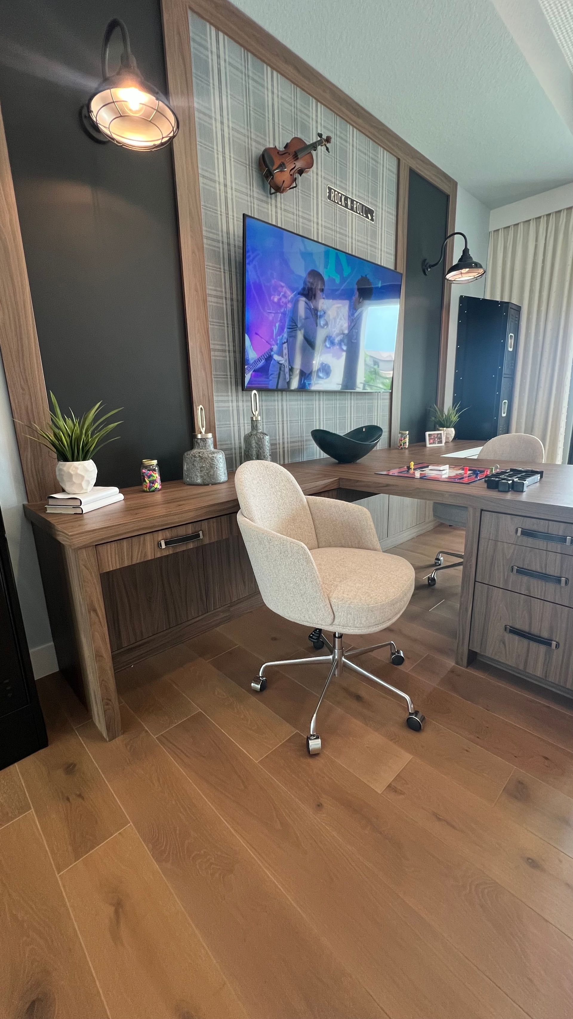 A bright home office with light wood floors, a desk with a white upholstered chair, and a wall-mounted TV.