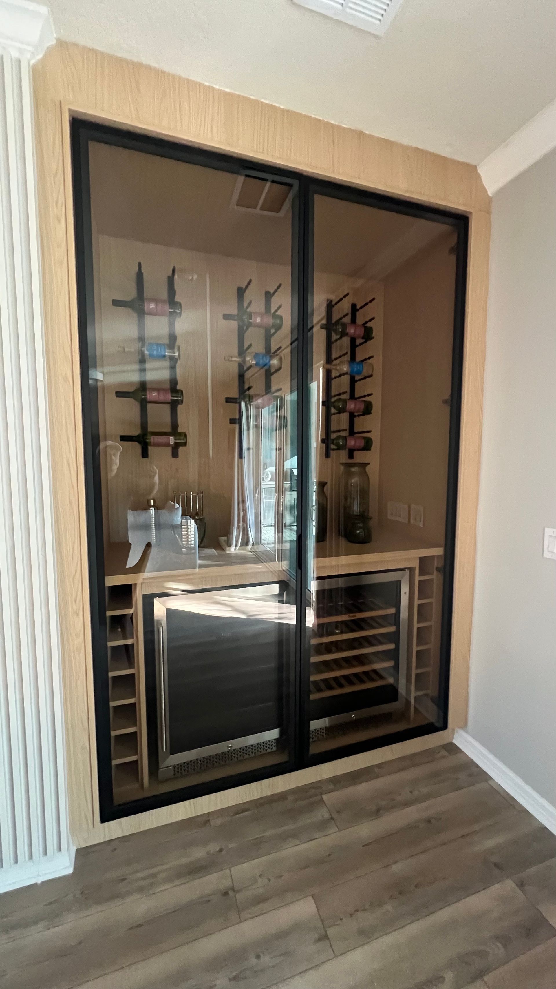 A modern wine display built into a light-wood wall, featuring glass doors, metal wall racks, and under-counter refrigerators.