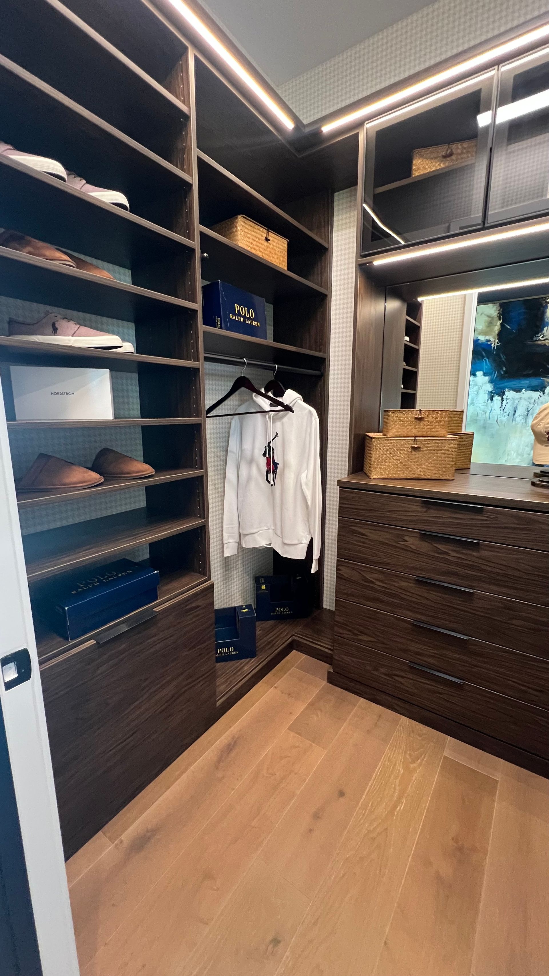 Walk-in closet with dark wood shelving, hanging clothes, built-in drawers, and light wood flooring under warm lighting.