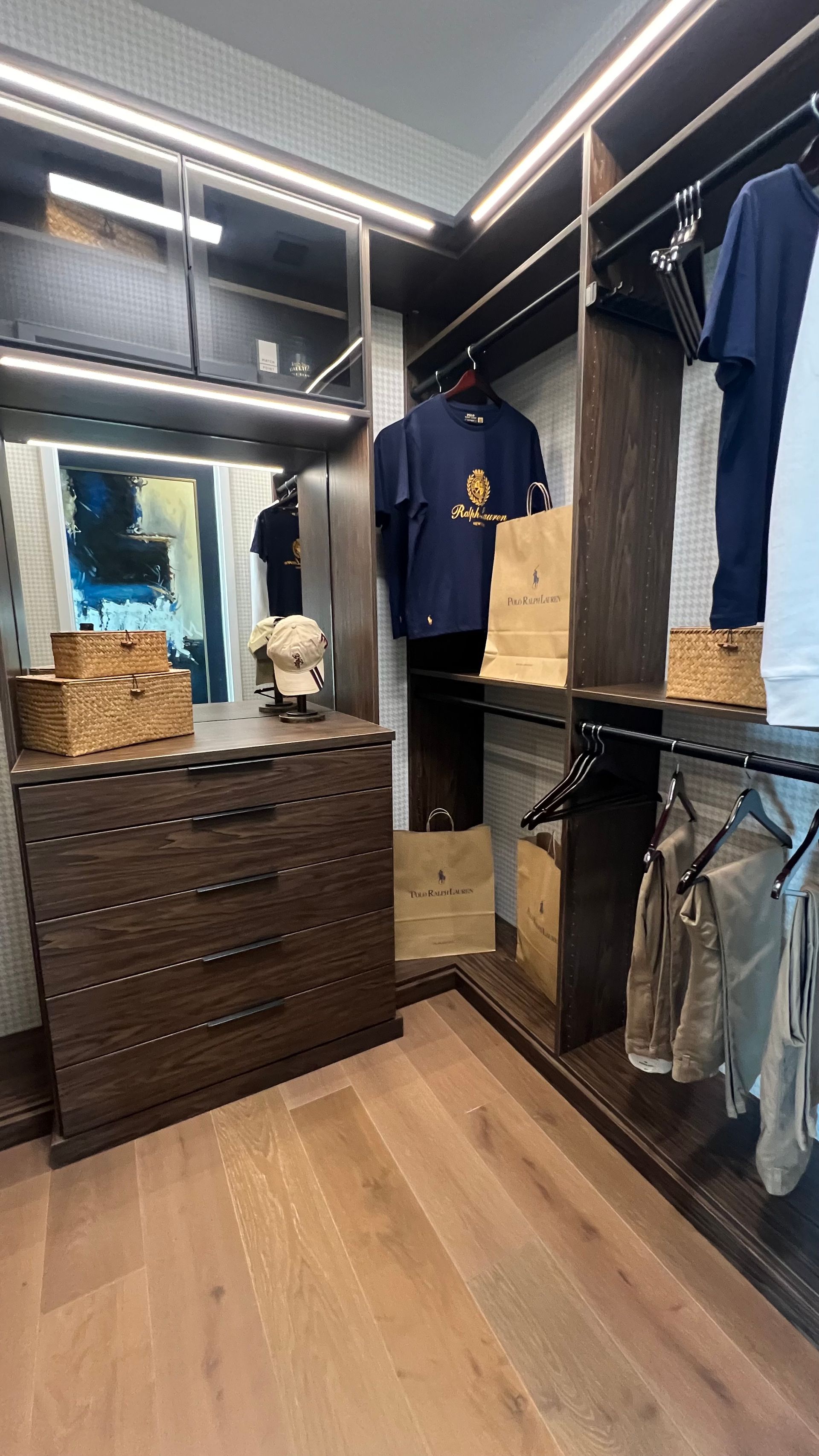 A modern walk-in closet with a wooden dresser, hanging clothes, integrated shelf lighting, and light wood flooring.