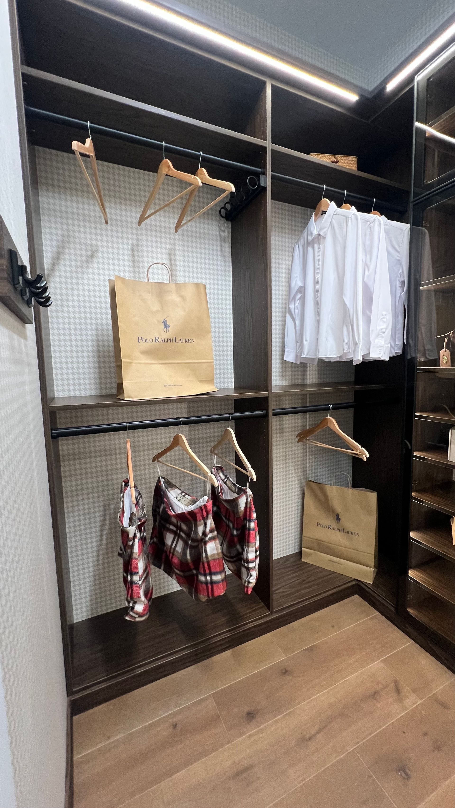 A modern walk-in closet featuring dark wood shelving, hanging clothes, shopping bags, and ambient LED lighting.