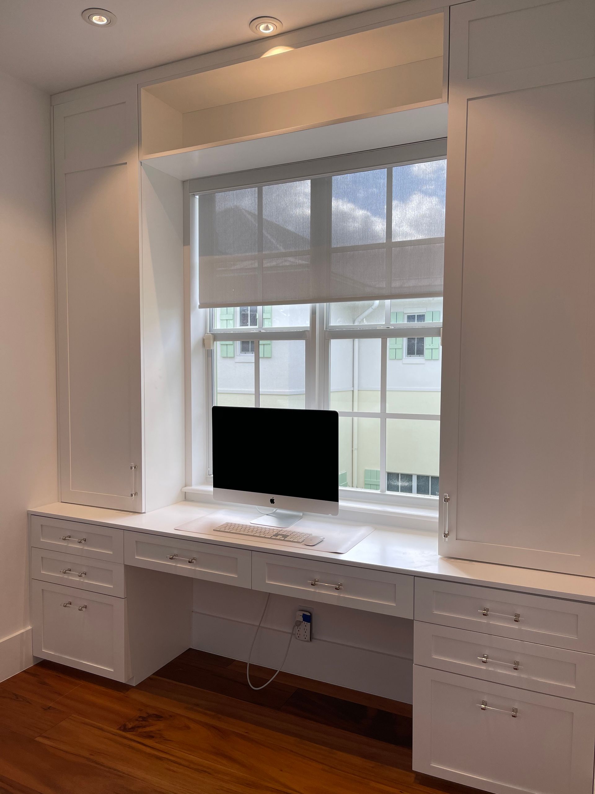 A white built-in desk unit with drawers and tall cabinets surrounding a window with a computer monitor on the desk.