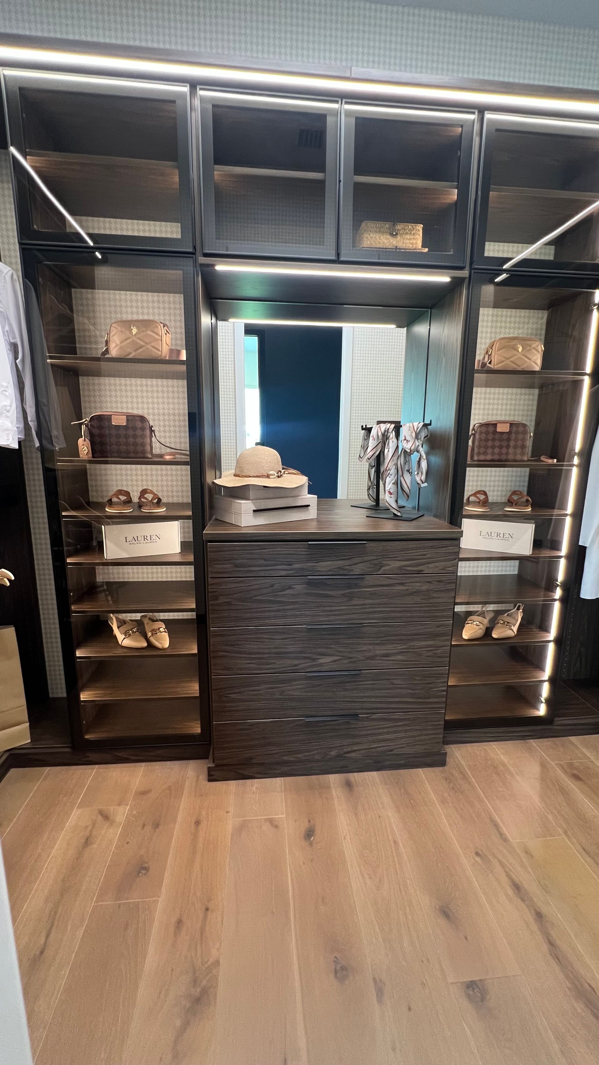 A custom dark wood closet system with open shelving, a central dresser, and integrated lighting in a room with wood floors.
