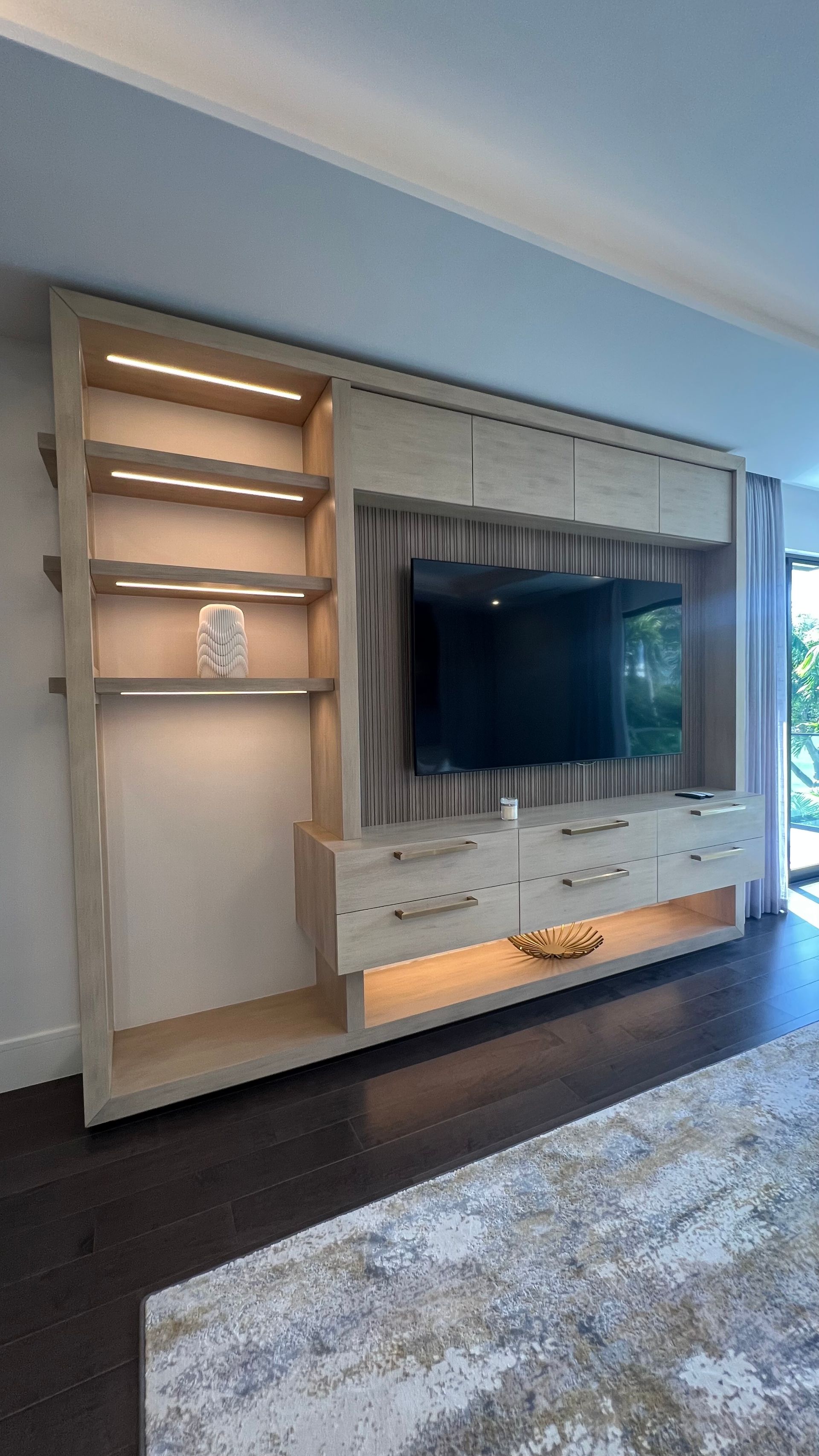 A light wood built-in media wall unit featuring shelves with LED lighting, a mounted TV, and lower storage drawers.