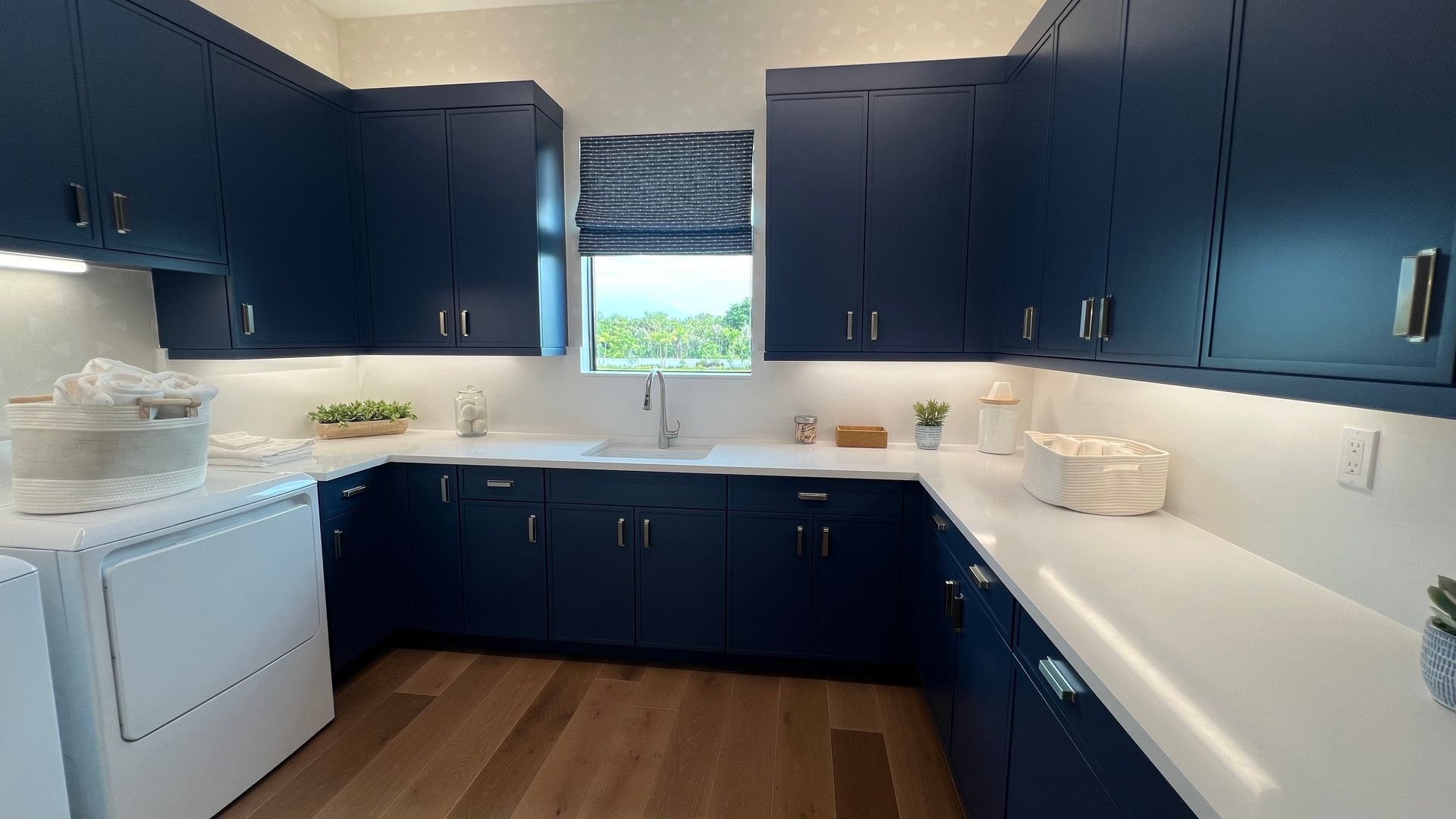 A laundry room with navy blue cabinets, white countertops, a large white sink, and light wood flooring.