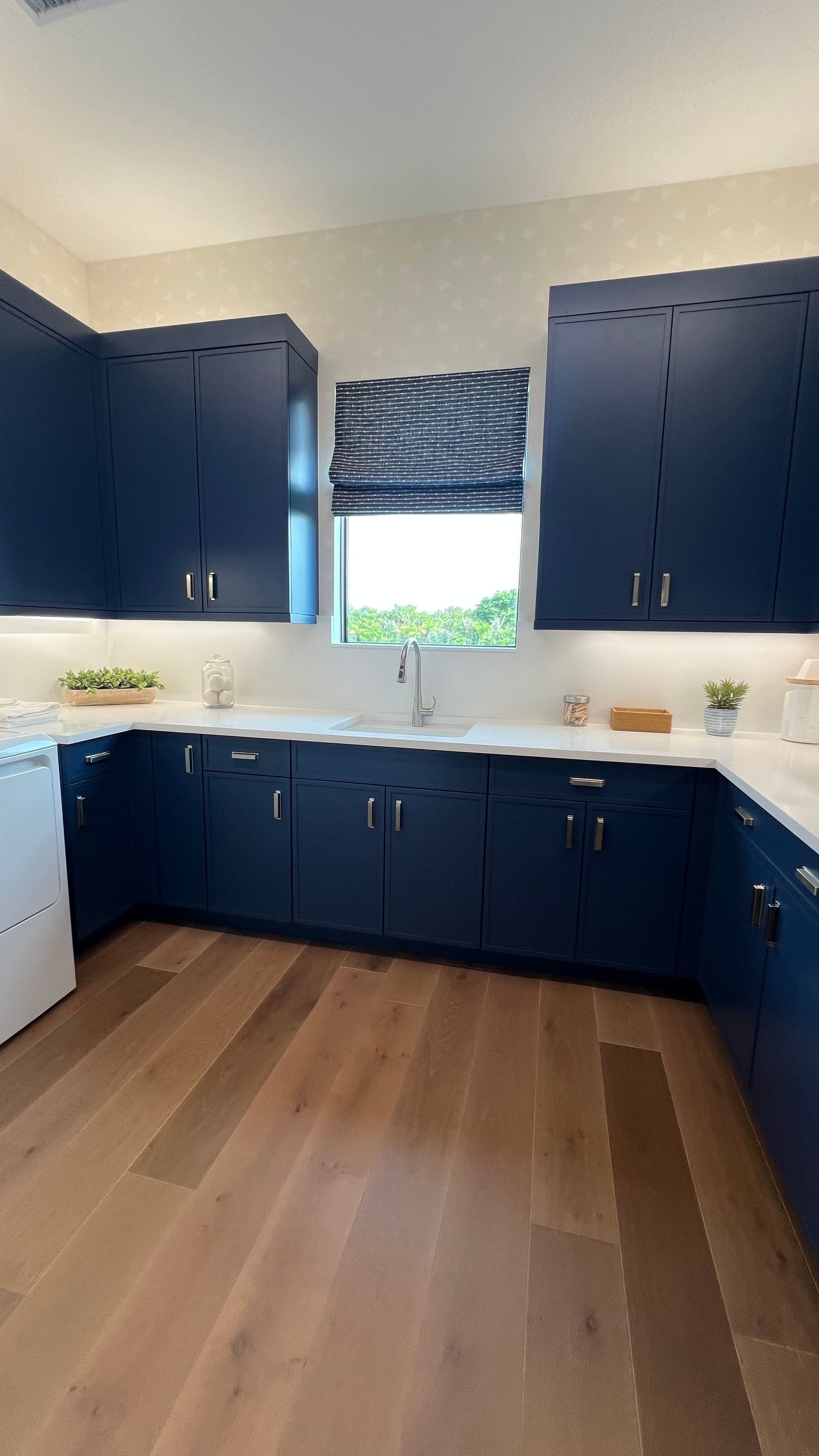 A kitchen featuring navy blue cabinets, white countertops, a central window with patterned shades, and light wood floors.