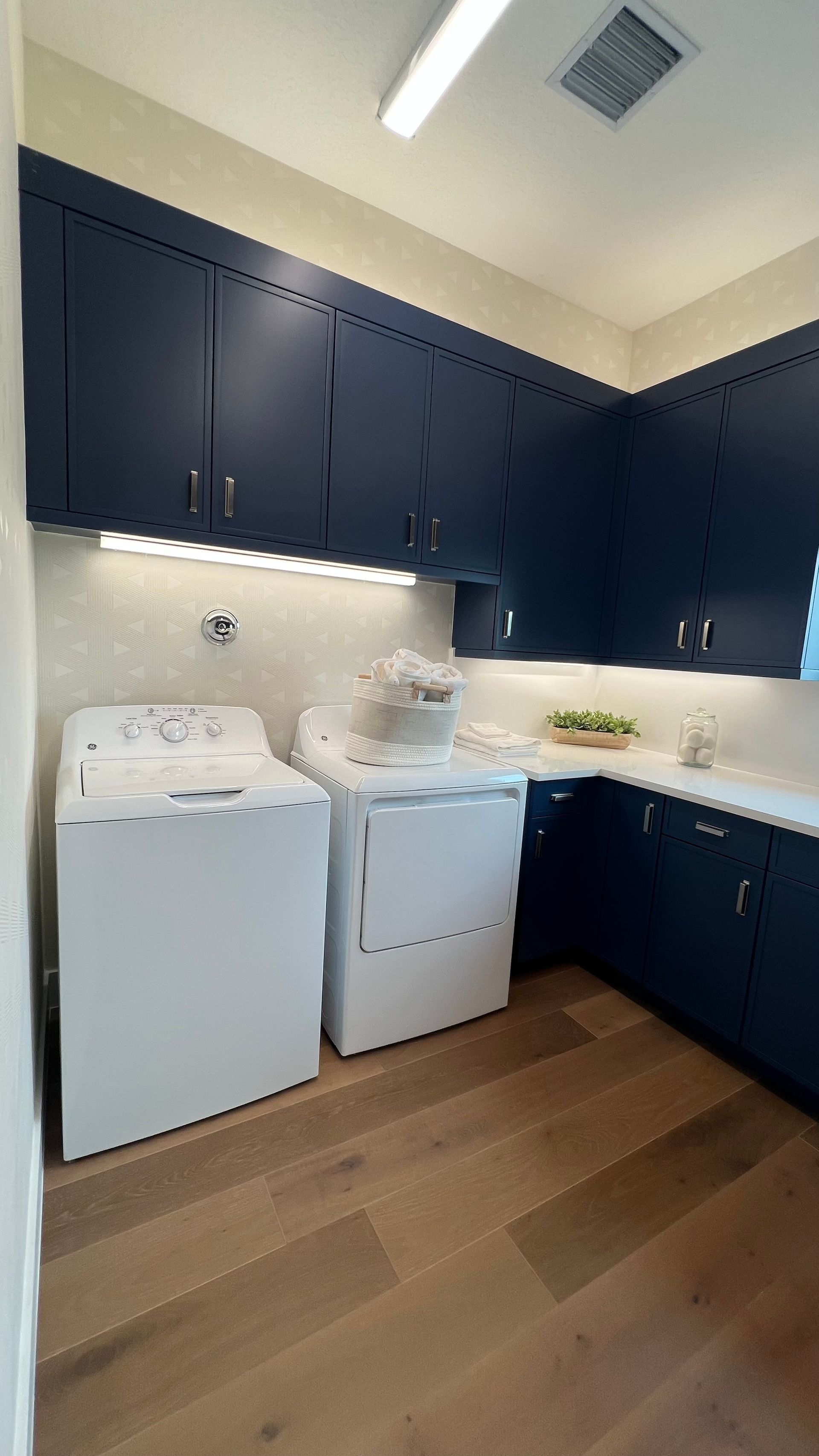 A laundry room with dark blue cabinets, white appliances, a light-colored countertop, and light wood flooring.