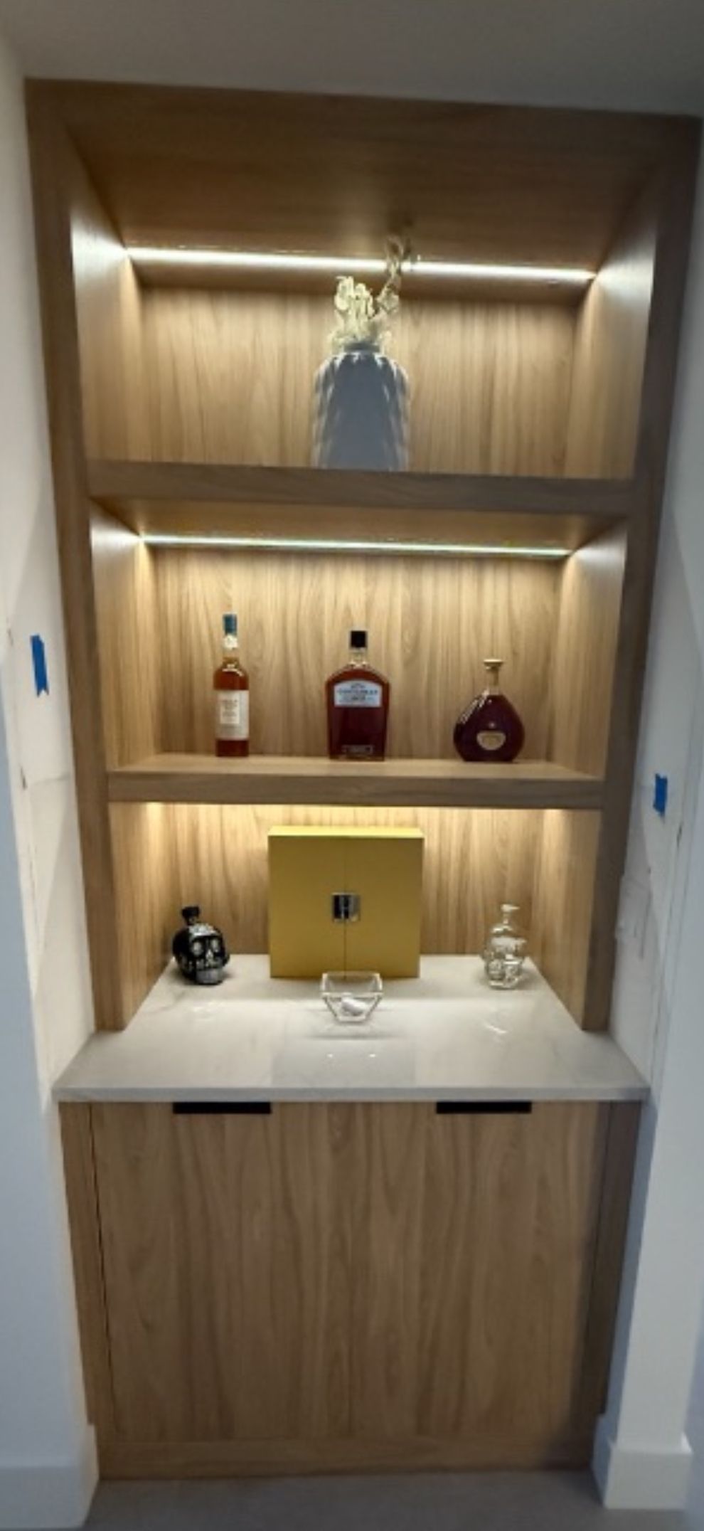 A light wood home bar built-in featuring illuminated shelves with bottles, a gold box, and a white marble counter.