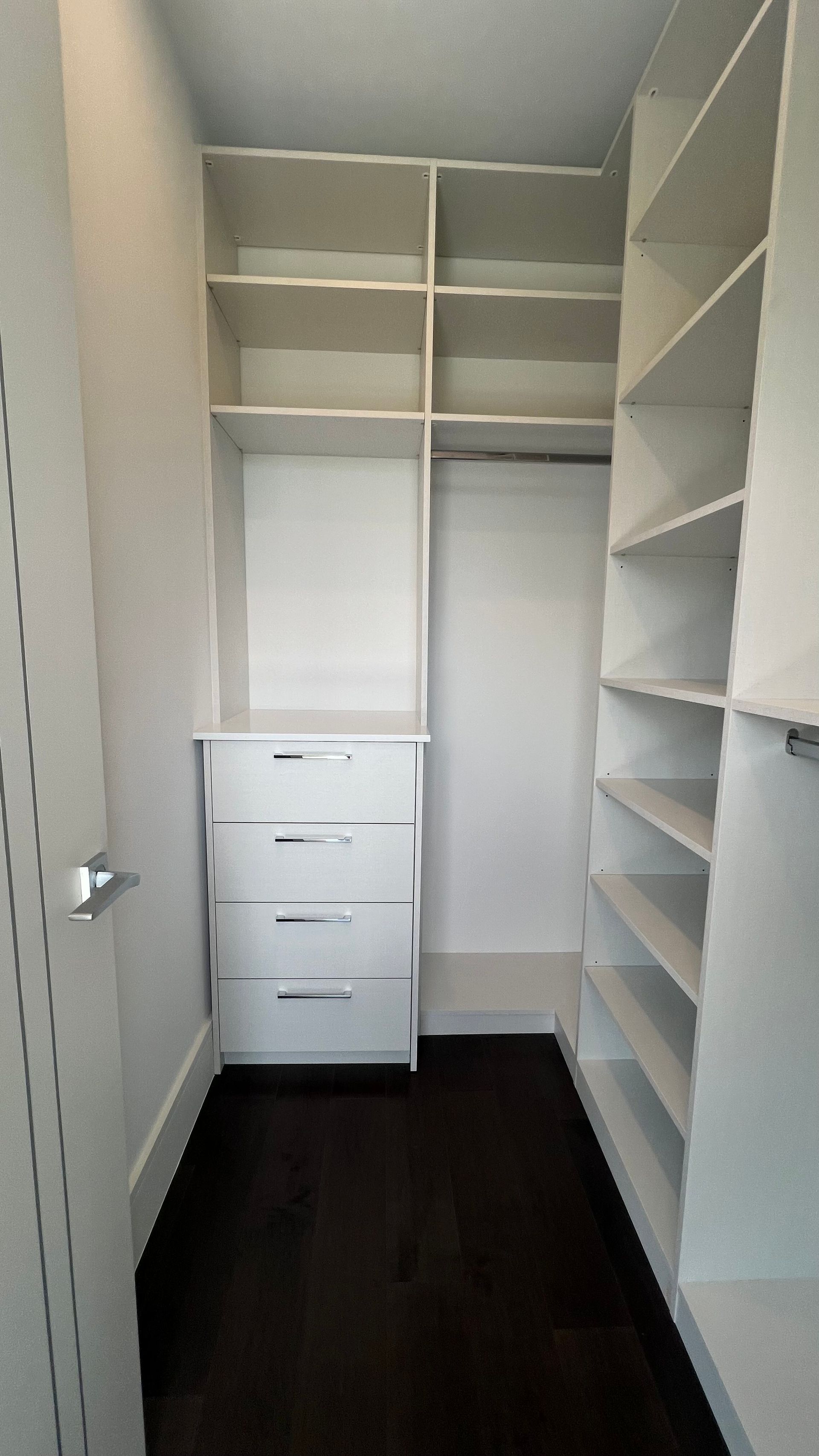 A walk-in closet featuring white wooden shelving, a set of three drawers, and dark hardwood floors.